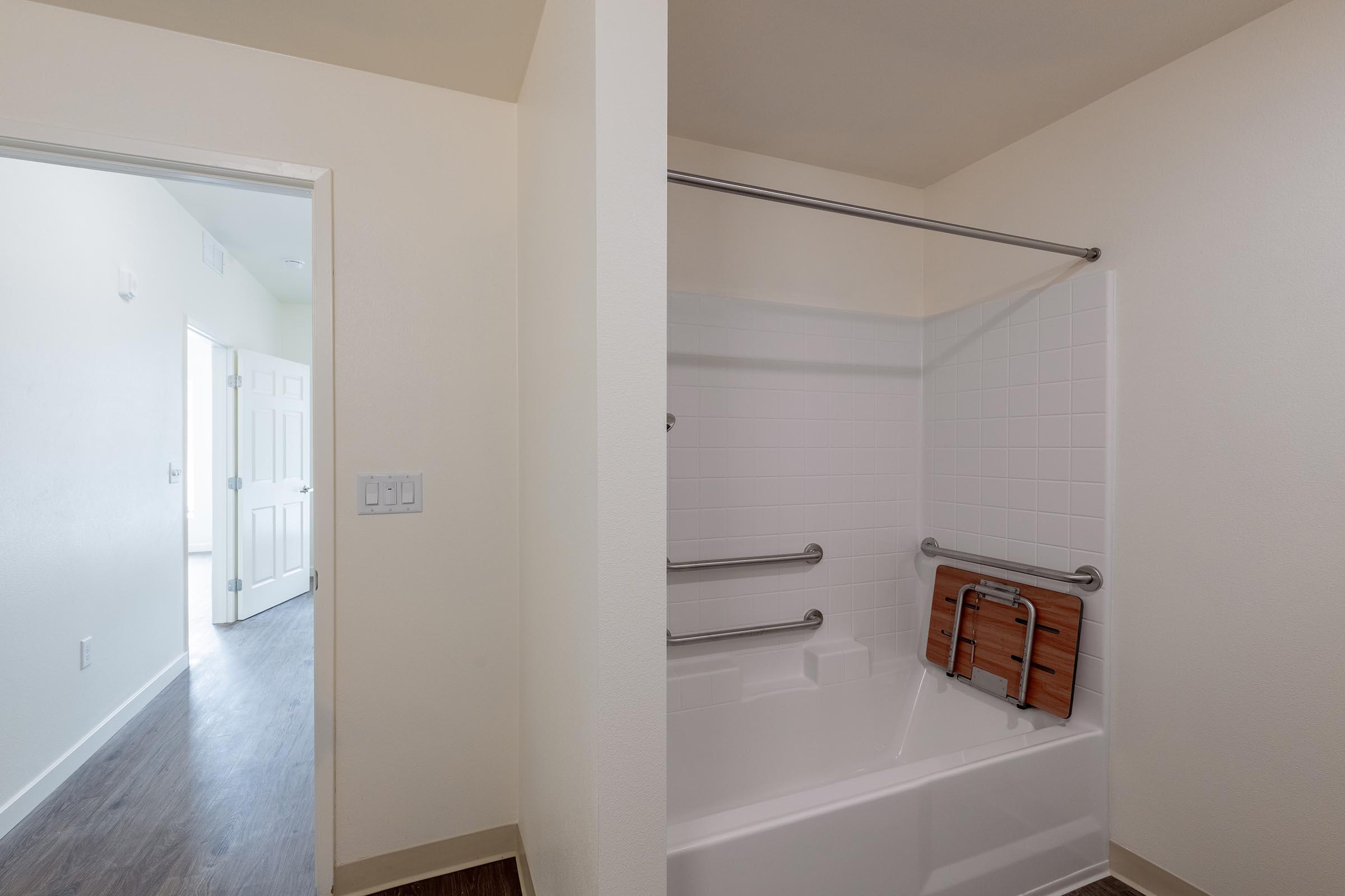 Interior view of a bathroom featuring a white bathtub with grab bars, an accessible shower curtain rod, and a wooden shower seat leaned against the tub. The wall is painted in a light color, and a doorway can be seen in the background leading to another room with closed doors.