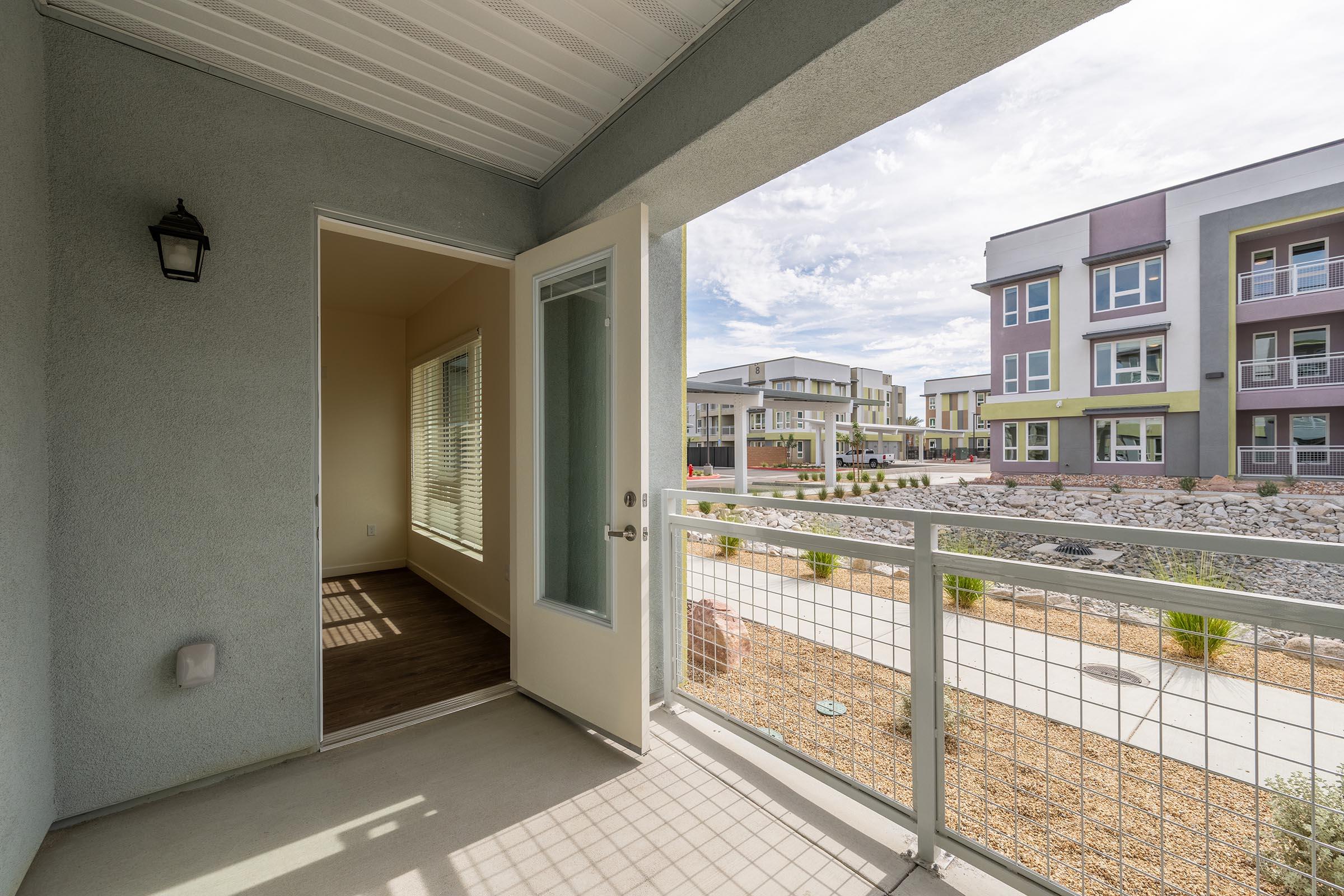 View from a balcony showing an entrance door leading inside, with a well-lit room visible through the open door. The exterior features modern apartment buildings in the background, landscaped areas with rocks and plants, under a partly cloudy sky.