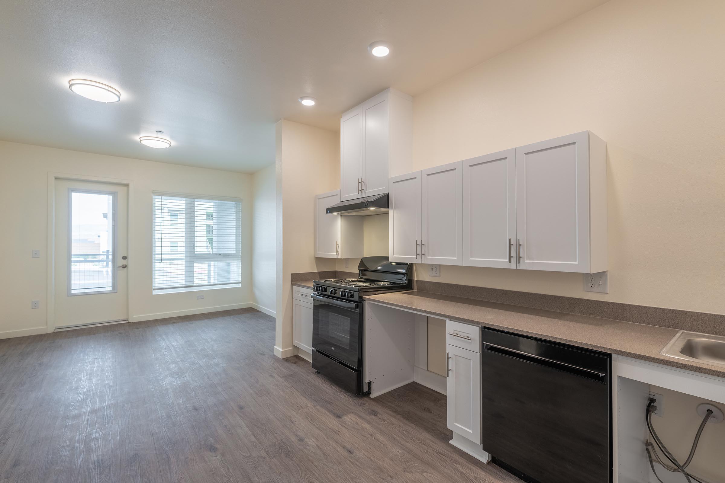 An interior view of a modern kitchen and dining area featuring white cabinetry, a black stove, and a stainless steel dishwasher. Large windows allow natural light into the space, complemented by recessed lighting. The floor is a light wood finish, and there’s a sliding glass door leading outside.
