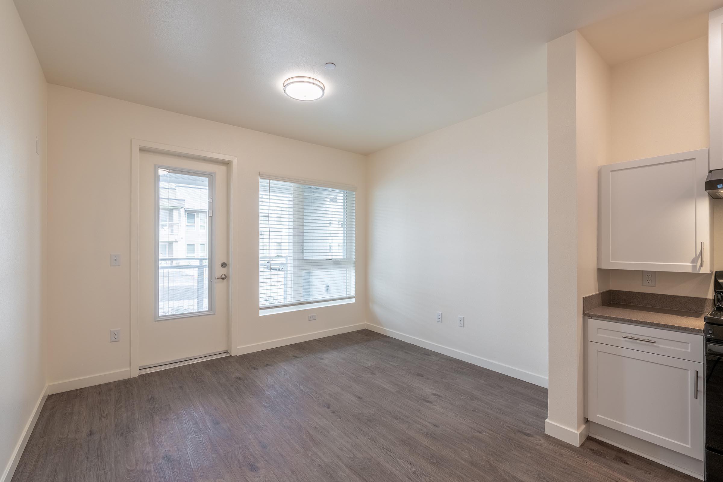 Empty interior of a room featuring a door leading outside, a window with blinds, and a small kitchenette with cabinets and a countertop. The walls are painted a light color, and the floor is a dark wood-like laminate, creating a modern and minimalist aesthetic.