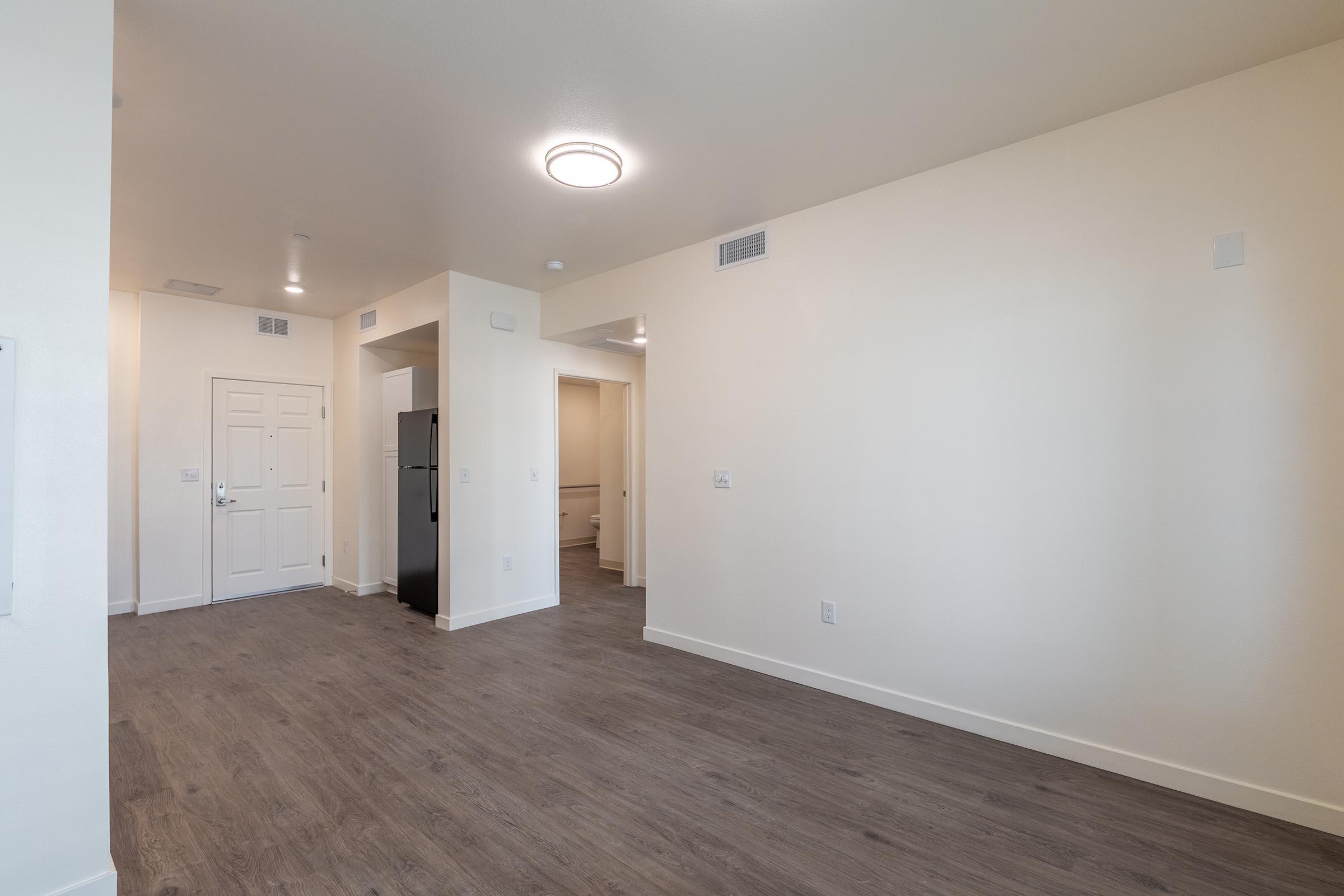 A spacious, empty room with light-colored walls and a wood-like laminate floor. The entrance is visible with a door on the left, a small hallway leading to a bathroom or additional room at the back, and a kitchen area with a refrigerator. The ceiling has recessed lighting fixtures.