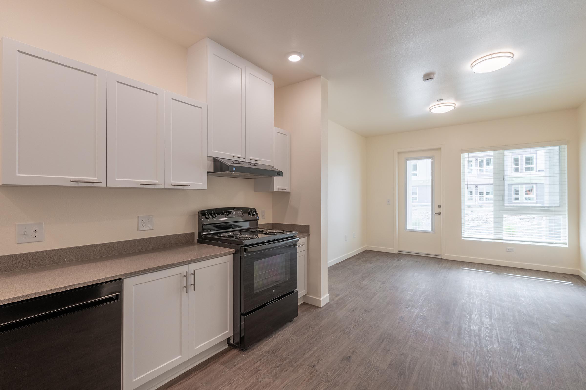 A modern kitchen featuring white cabinets, a black stove, and a stainless steel dishwasher. The space has a spacious layout with natural light coming from large windows. A door leads to an outdoor area, complemented by neutral-colored walls and a stylish laminate floor.