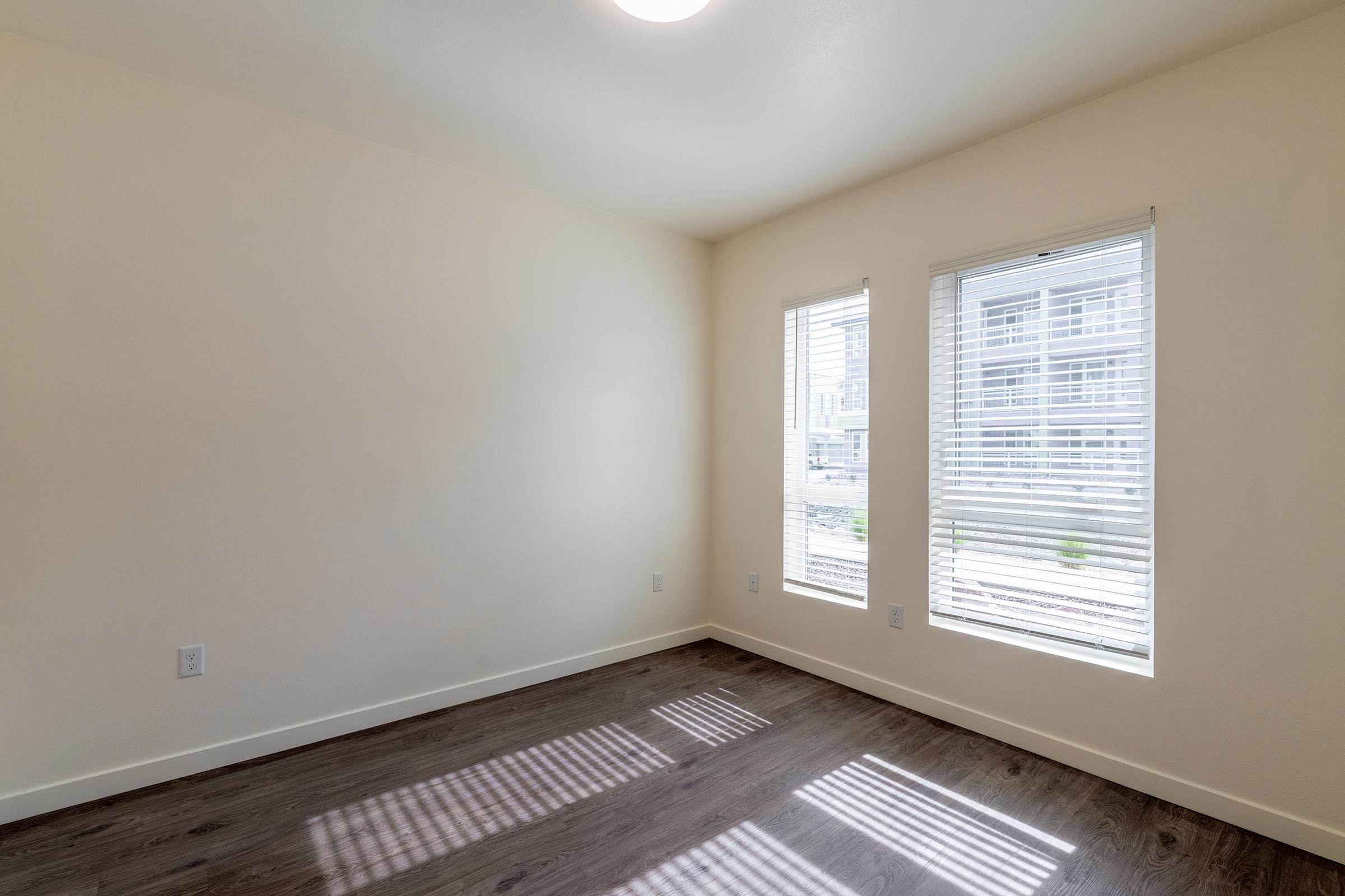 Empty room with light-colored walls and two windows with white blinds. Natural light streams in, casting shadows on the brown wooden floor. The space is unadorned, creating a minimalist and airy atmosphere. No furniture or decor is visible.