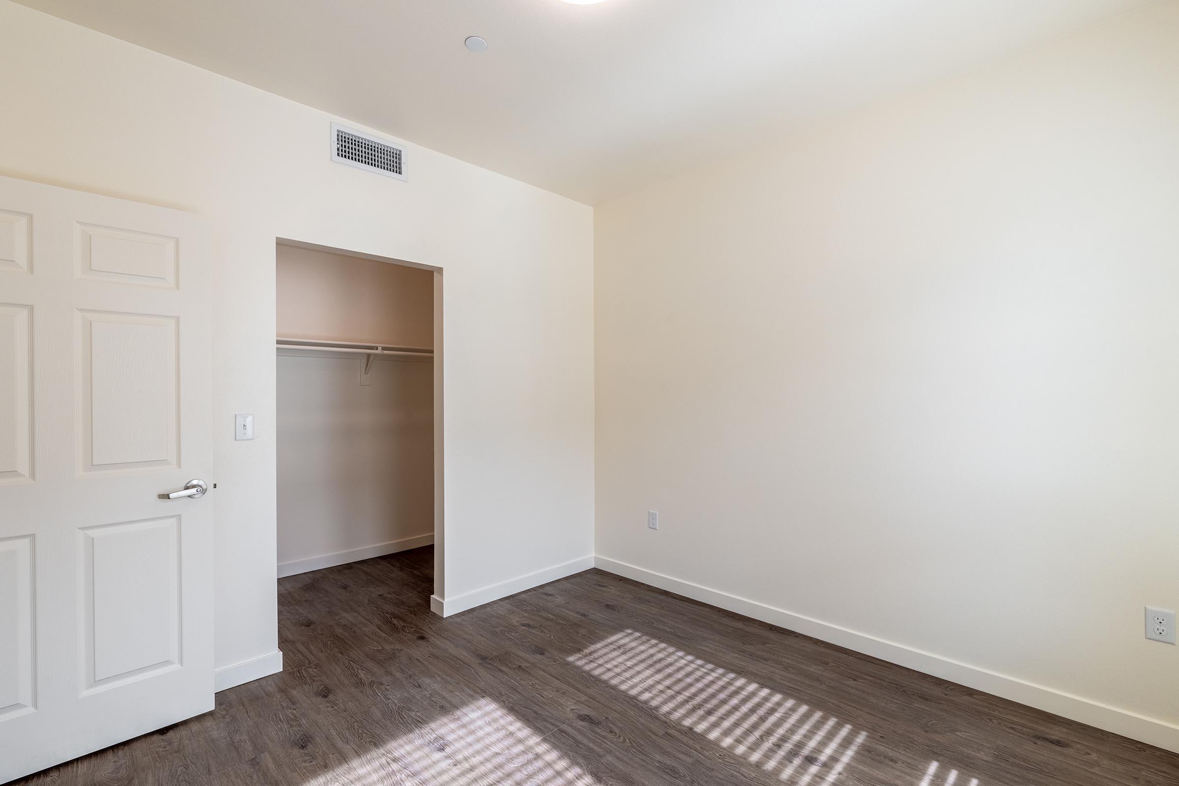 A clean, empty room with light-colored walls and wood-like flooring. There's a closet with a white door and a single shelf visible inside. The room has natural light streaming in, casting shadows on the floor. No furniture or decorations are present, creating a spacious and minimalistic atmosphere.