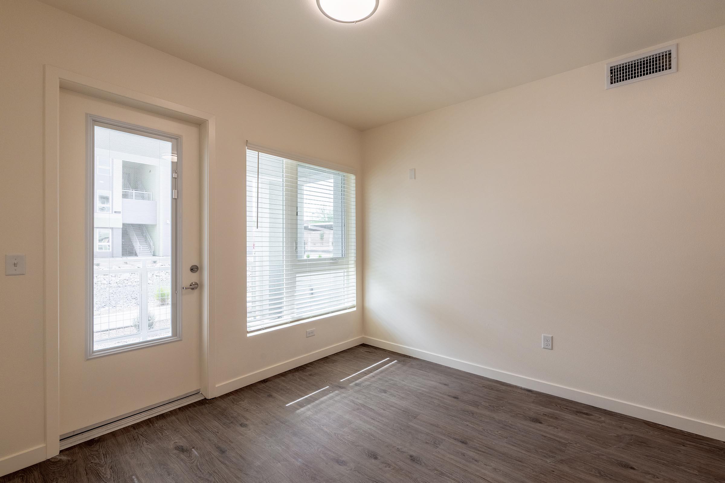 A bright, empty room featuring a large window with blinds, a door leading outside, and natural wood flooring. The walls are painted a light color, creating an open and airy atmosphere. The overall design is modern and minimalistic, with a focus on natural light.