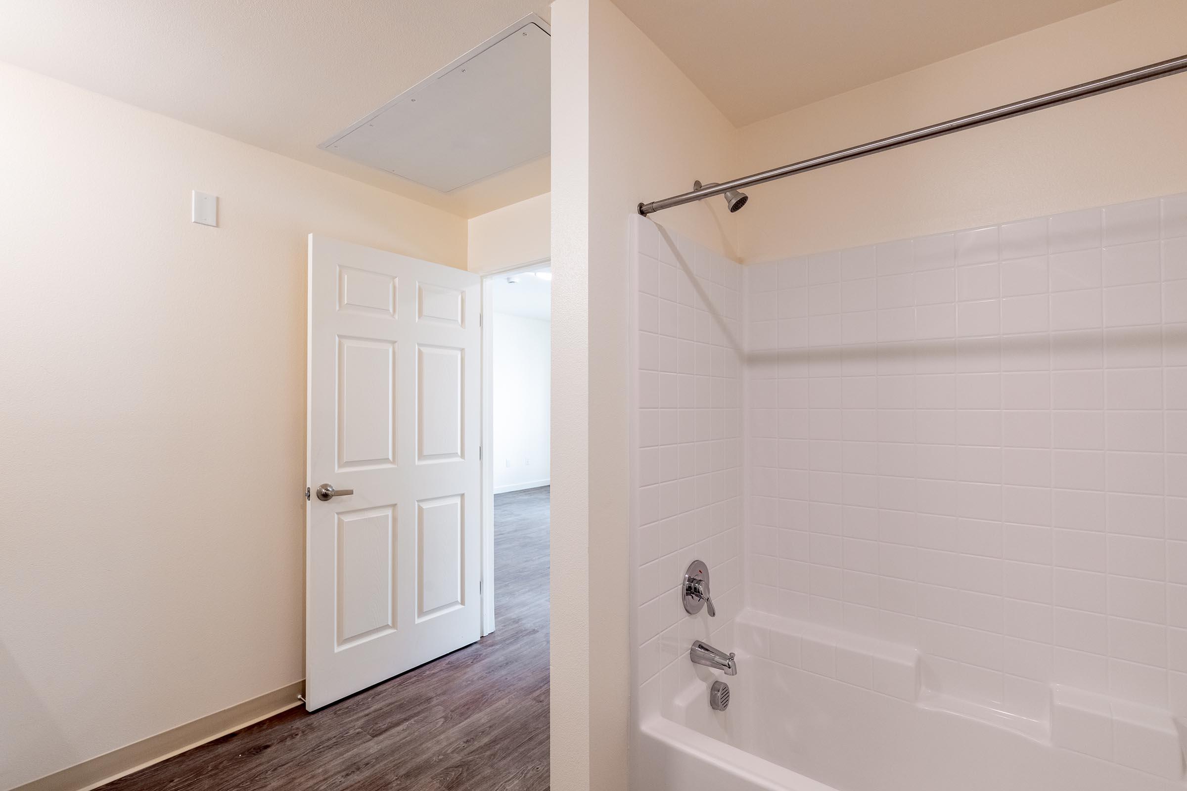 Interior of a bathroom showing a bathtub with a showerhead, a tiled wall, and a silver grab bar. A partially opened door leads to another room with wooden flooring. The walls are painted a light color, creating a spacious and clean atmosphere.
