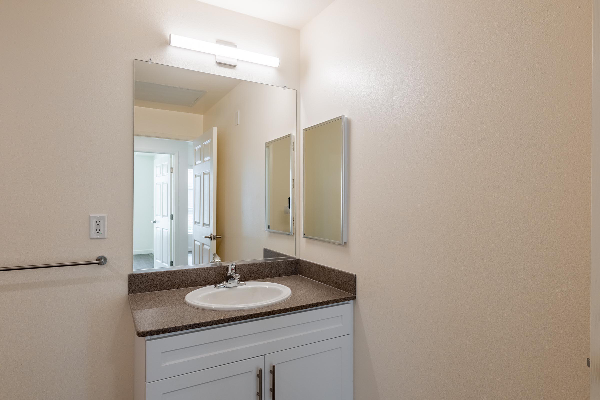 A clean bathroom with a light-colored wall, featuring a simple white vanity with a sink, a large mirror, and two smaller mirrors on either side. The countertop is a dark color, and the lighting provides a bright and airy feel to the space. A doorway is visible in the background, leading to another room.