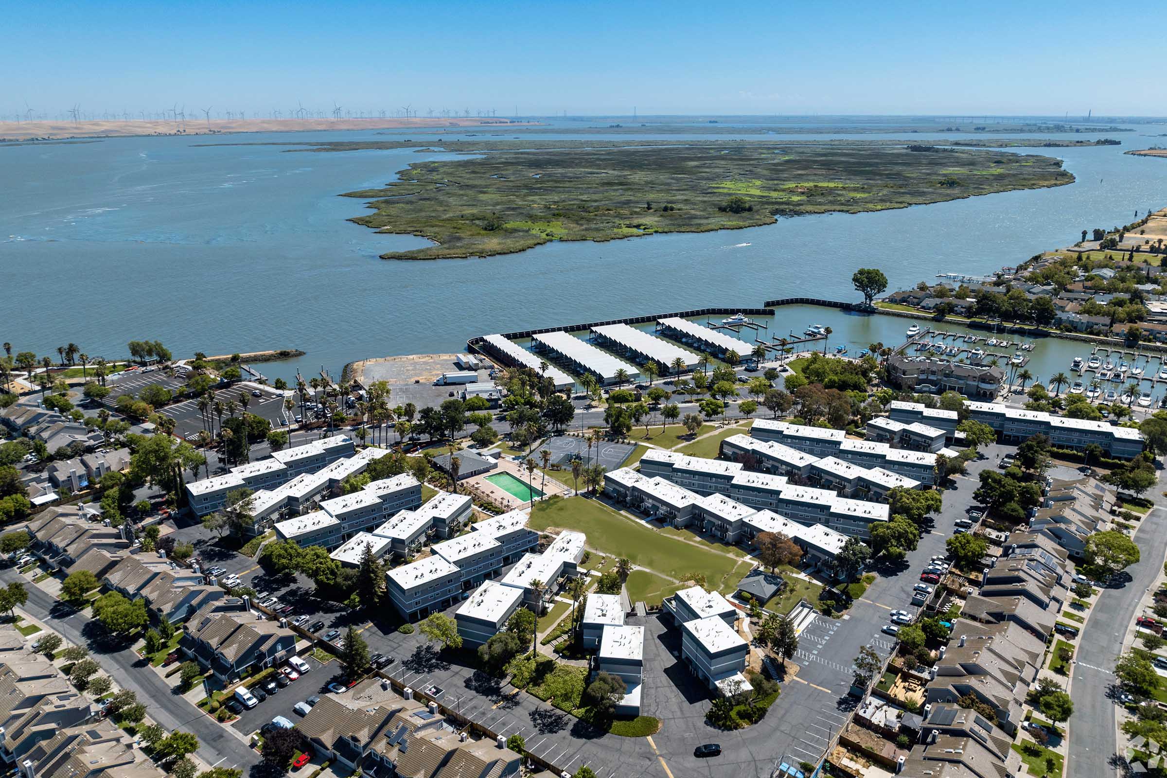 Aerial view of a waterfront residential area featuring multiple buildings arranged around green spaces and a water body. In the background, a marina with boats and a natural landscape, including an island and distant wind turbines, is visible under a clear blue sky.