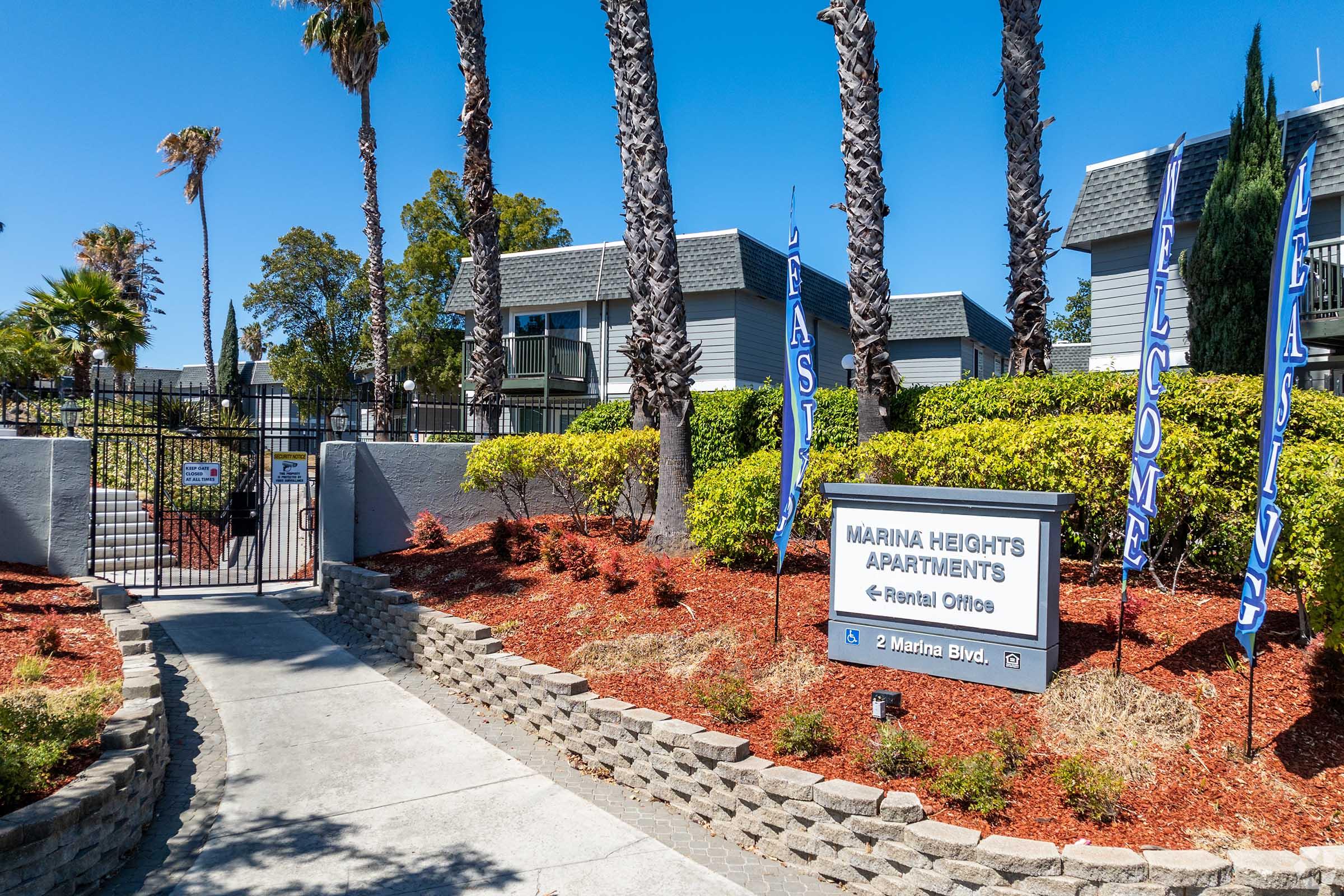 Gate entrance to Marina Heights Apartments, featuring landscaped pathways with palm trees and colorful flags. A sign indicates the rental offices, with buildings and greenery in the background under a clear blue sky.