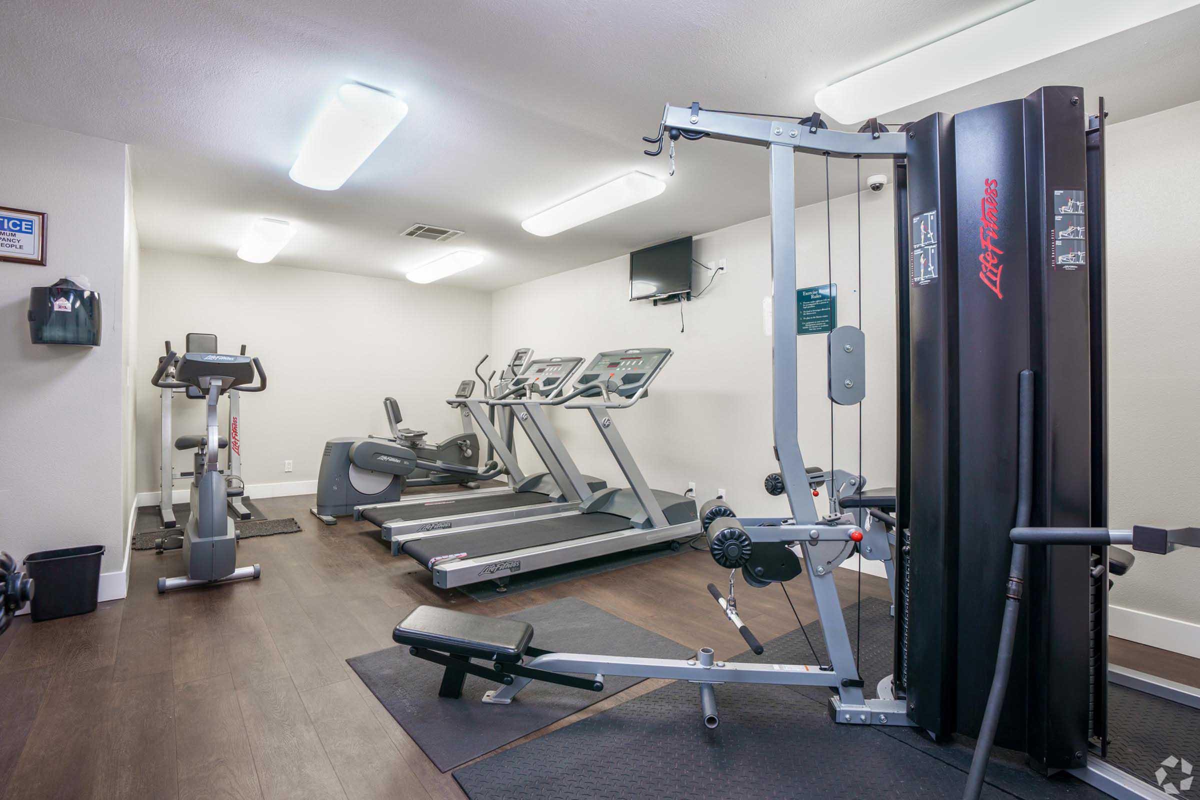A modern gym interior featuring exercise equipment. There are two treadmills and an elliptical machine against one wall, while a multi-gym machine is positioned nearby. The flooring is dark wood, and there are bright overhead lights illuminating the space, creating a clean and inviting workout environment.