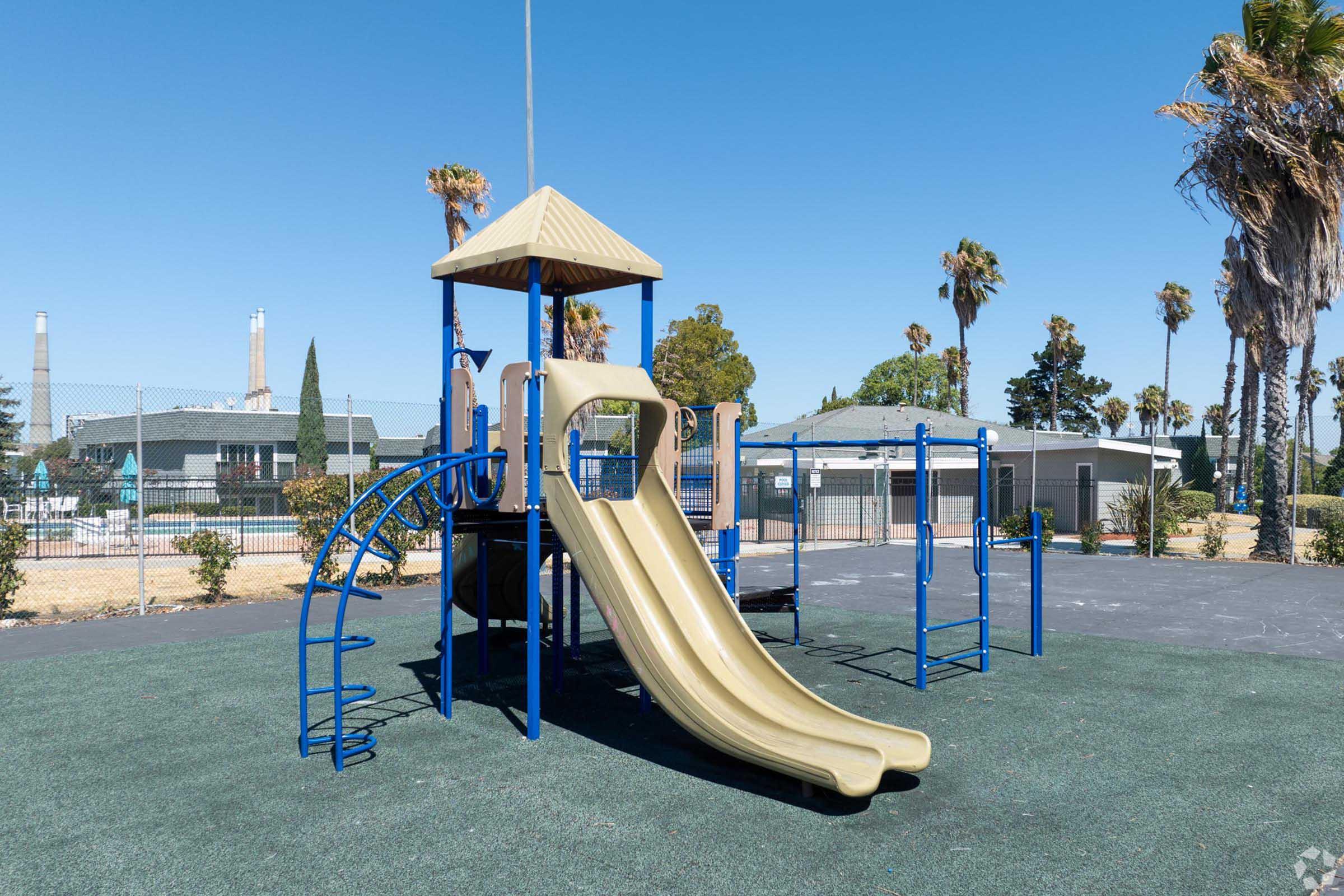 Playground equipment featuring a tall slide and a play tower with a canopy. The structure is blue and tan, situated on a grassy surface. Palm trees and a building are visible in the background against a clear blue sky.
