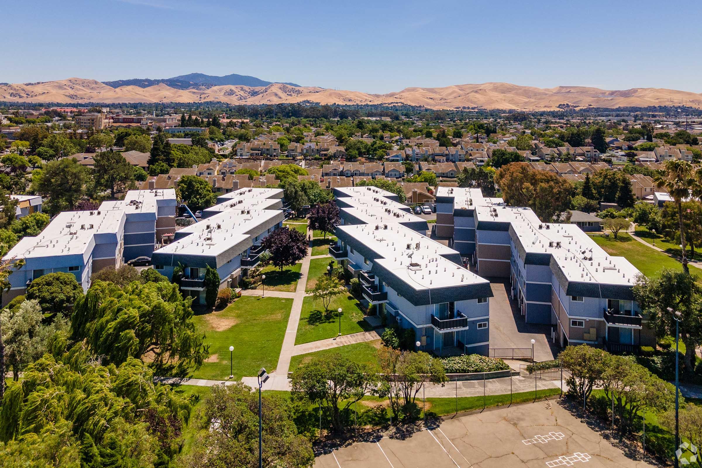 Aerial view of a residential area featuring multiple apartment buildings arranged around green lawns and pathways. In the background, rolling hills are visible under a clear blue sky, with a mix of urban and natural landscapes. The scene suggests a suburban environment with ample greenery.