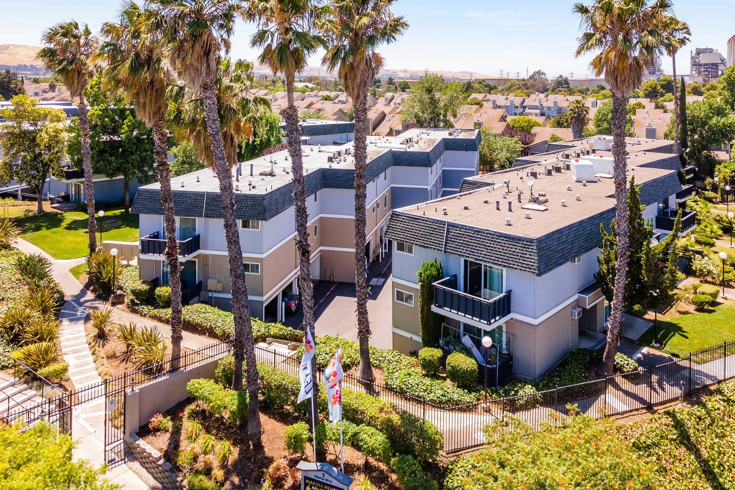 Aerial view of a residential complex featuring multiple interconnected buildings surrounded by palm trees and landscaped greenery. Pathways and decorative flags can be seen in the vicinity, indicating a welcoming environment. Clear blue skies and sunny weather enhance the outdoor setting.