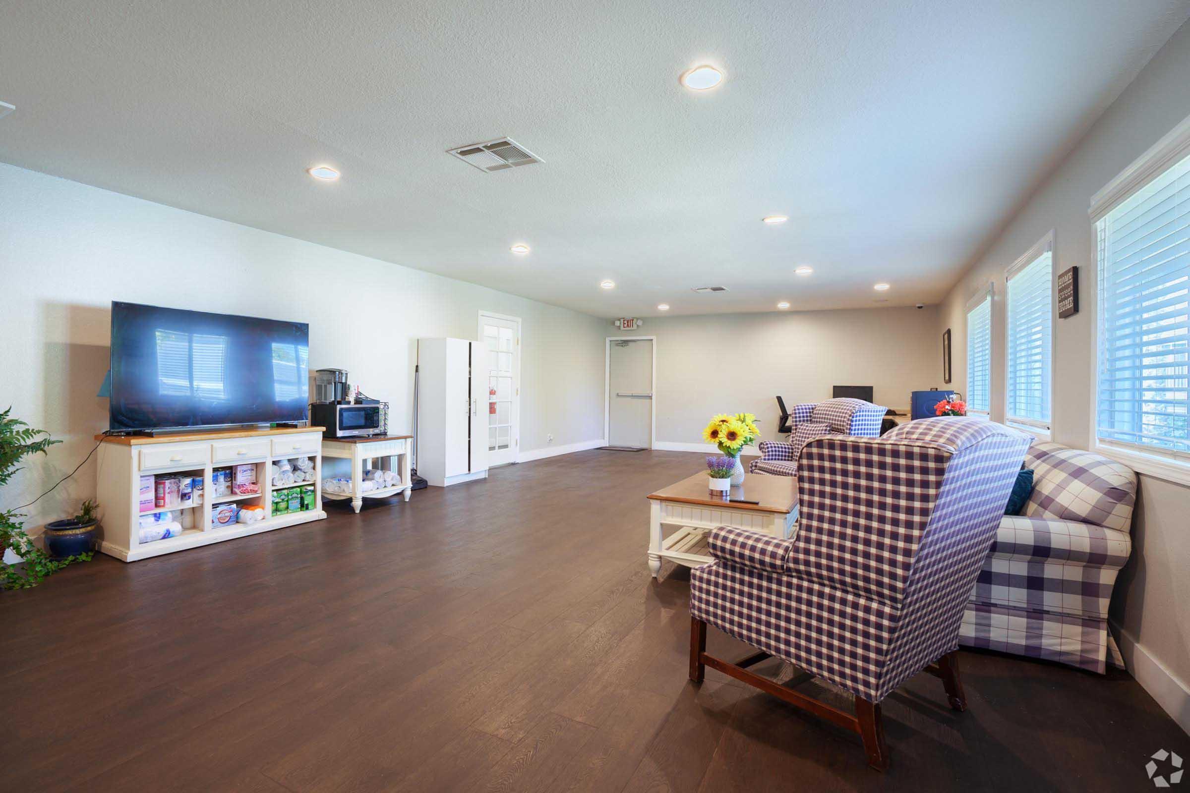 A spacious and well-lit living area featuring wooden flooring, two patterned armchairs, and a cozy sofa. A large flat-screen TV is mounted on the wall, and there's a white cabinet with various items. Sunlight streams through several windows, while a small table with sunflowers adds a touch of decor.