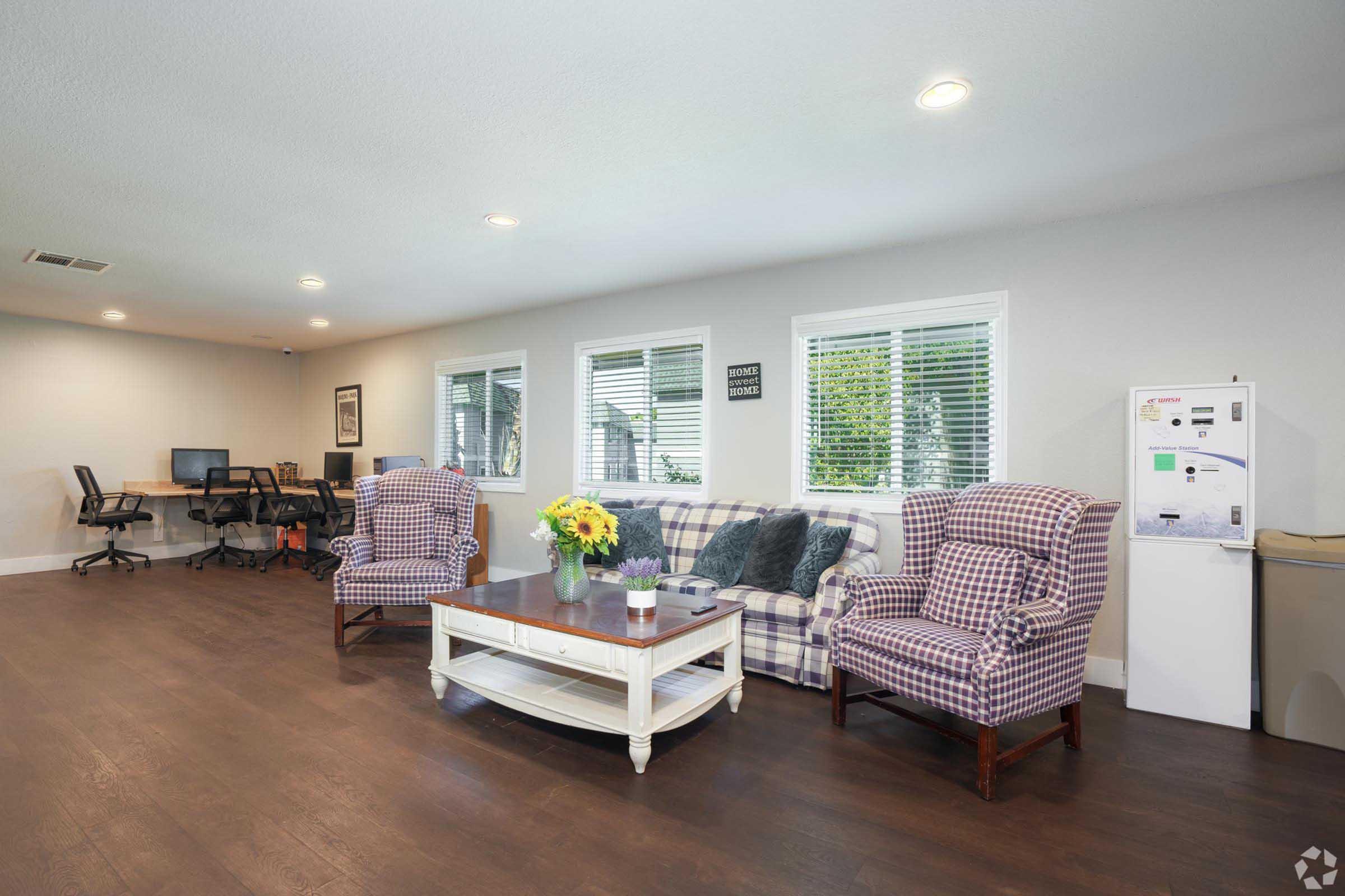 A spacious indoor lounge area featuring two plaid armchairs and a couch, a coffee table with a vase of flowers, and a small office setup with two desks and computers. Large windows allow natural light, and a beverage machine is visible in the corner.