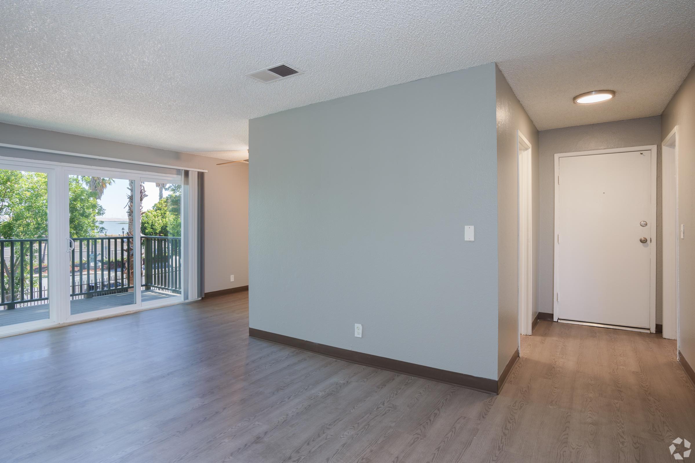 Interior view of an empty room featuring light gray walls and laminate flooring. There is a doorway leading to an outdoor balcony with green trees visible outside. The entrance is seen on the right, and natural light is coming in from the balcony door. The space has a modern, minimalist design.