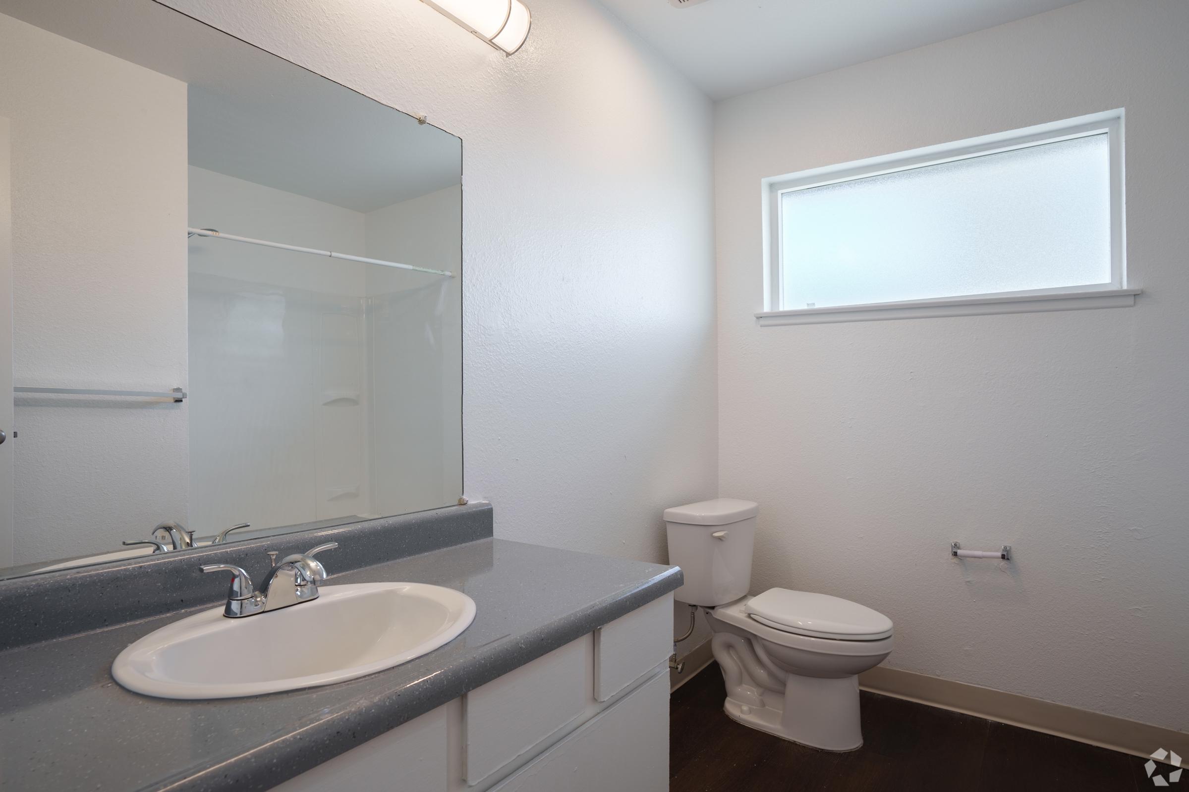 A clean, modern bathroom featuring a single sink with a faucet, a toilet, and a glass shower enclosure. The walls are painted white, and there is a small window allowing natural light to enter the space. The floor is dark, contrasting with the light-colored fixtures and walls.