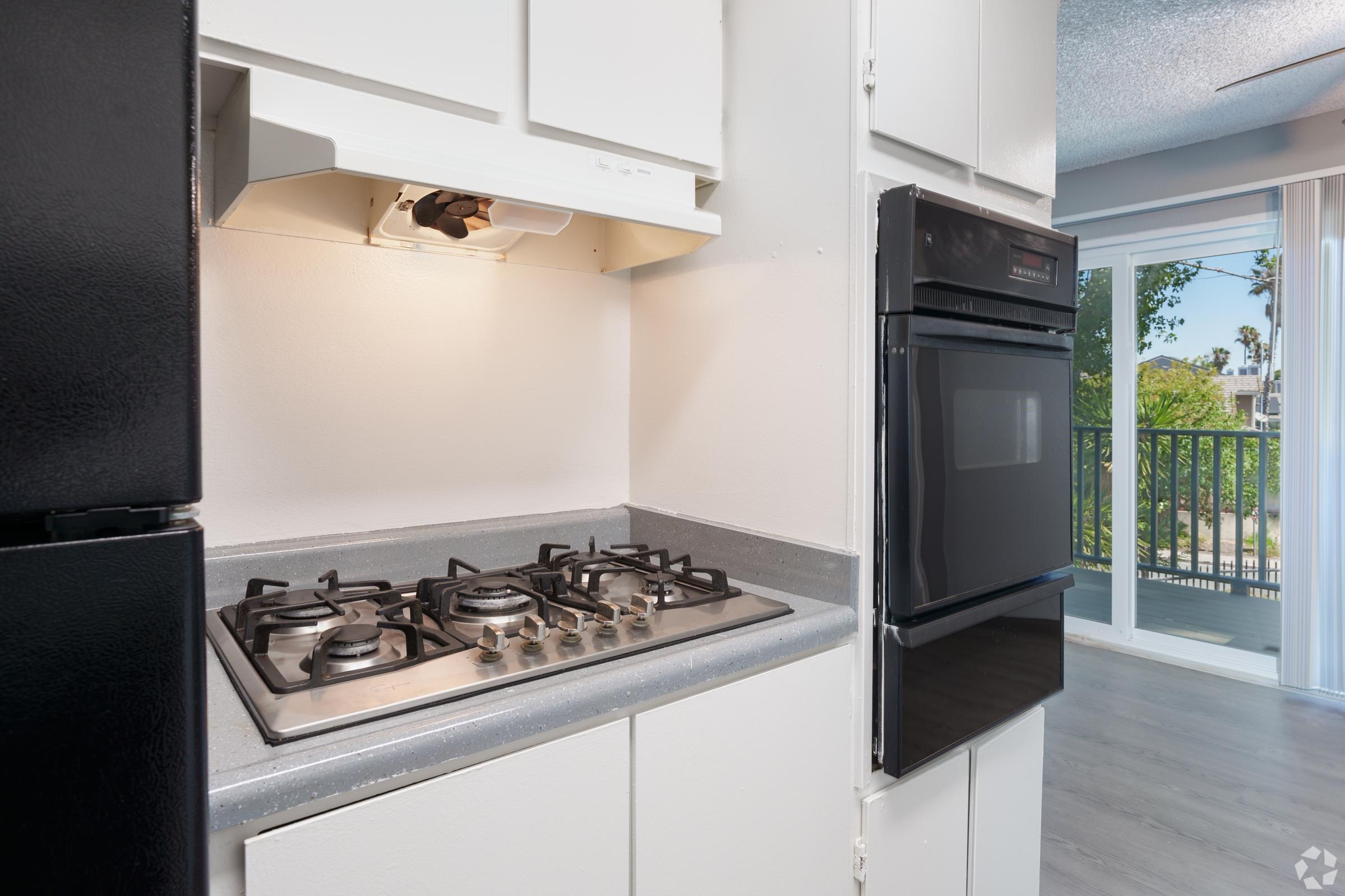 A modern kitchen with a gas stove, overhead exhaust fan, and black oven. The cabinets are white, and the countertop is gray. A window shows a view of an outdoor balcony with greenery, allowing natural light into the space.