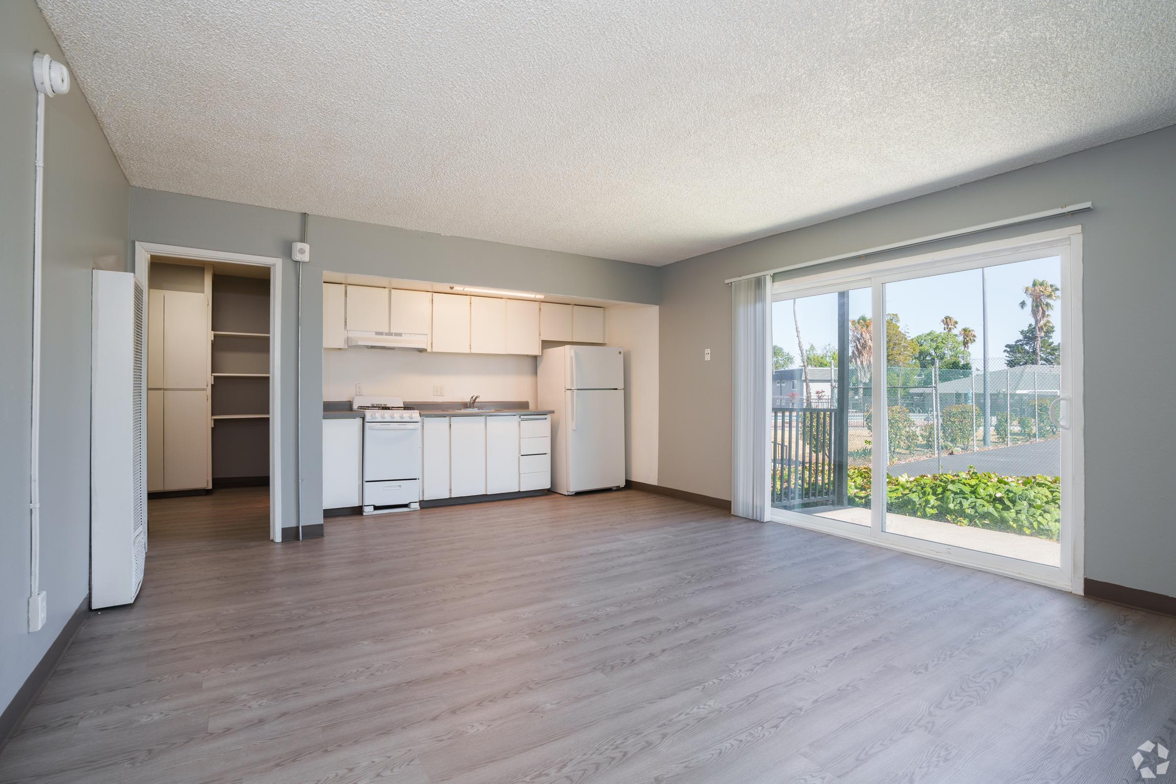A bright, minimalist living space featuring an open kitchen with white cabinets, a stove, and a refrigerator. The room has light wood flooring and large sliding glass doors leading to a patio with greenery. Walls are painted in neutral tones, creating a spacious and inviting atmosphere.