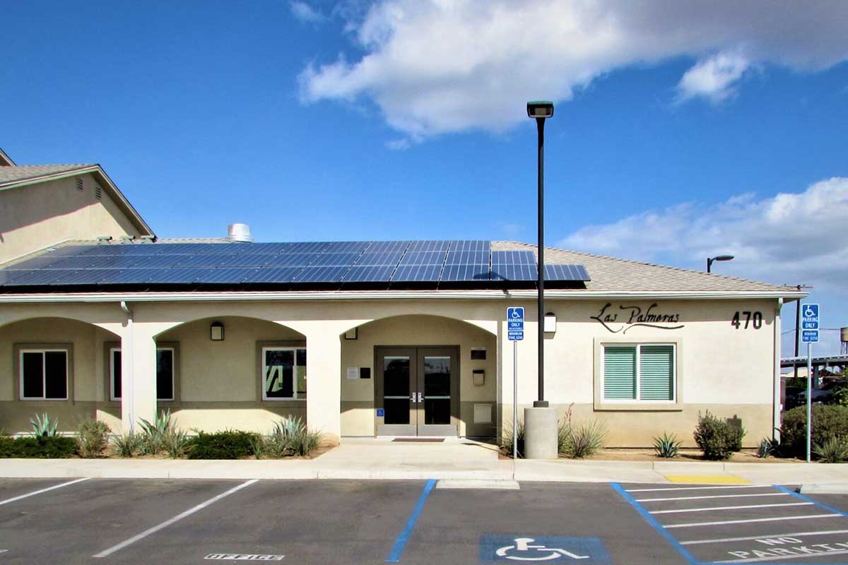 A single-story building with solar panels on the roof, featuring the name "Las Palmeras" above the entrance.