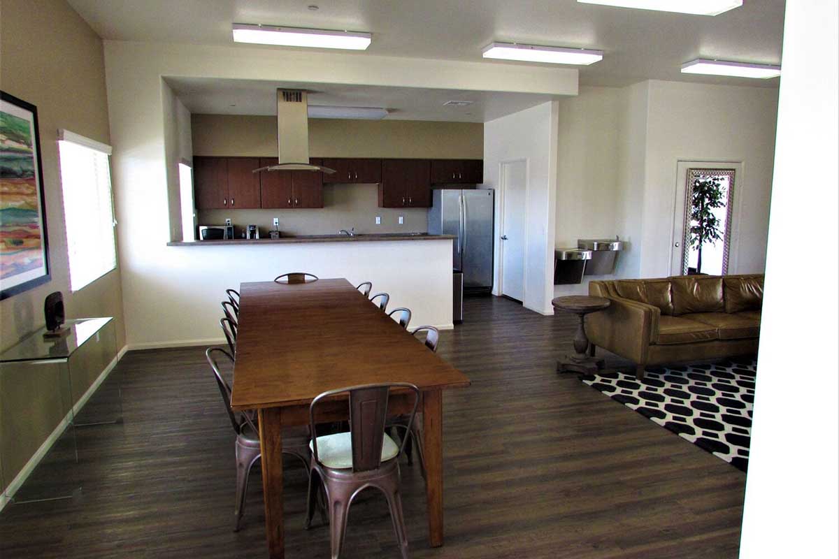 A spacious and modern living area featuring a long wooden dining table with metal chairs, a brown leather sofa, and a patterned rug.