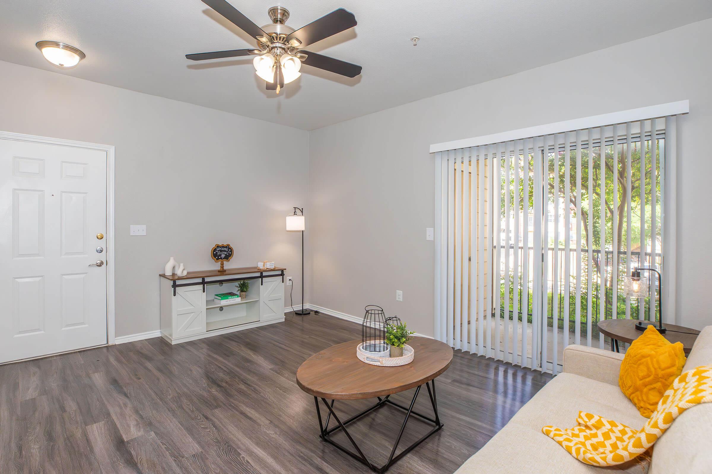 A cozy living room featuring a ceiling fan, light wood flooring, and a large window with vertical blinds. There's a beige couch adorned with a bright yellow pillow, a round coffee table in the center, and a small console table with decorative items. The space is well-lit and has a welcoming ambiance.