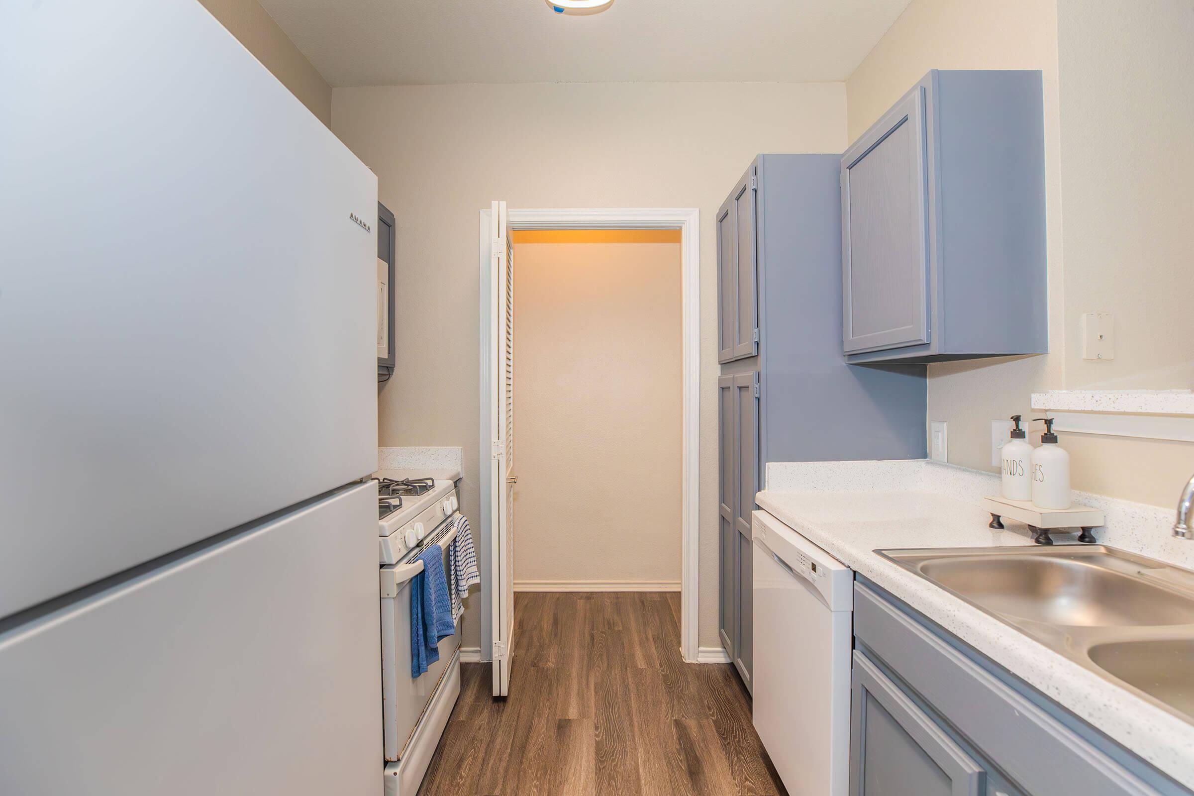 A modern kitchen featuring light blue cabinetry, a white countertop, and stainless steel appliances. The refrigerator is on the left, with a gas stove beside it. A doorway at the back leads to another space, while a sink and dishwasher are positioned on the right. The flooring is a warm wood tone.