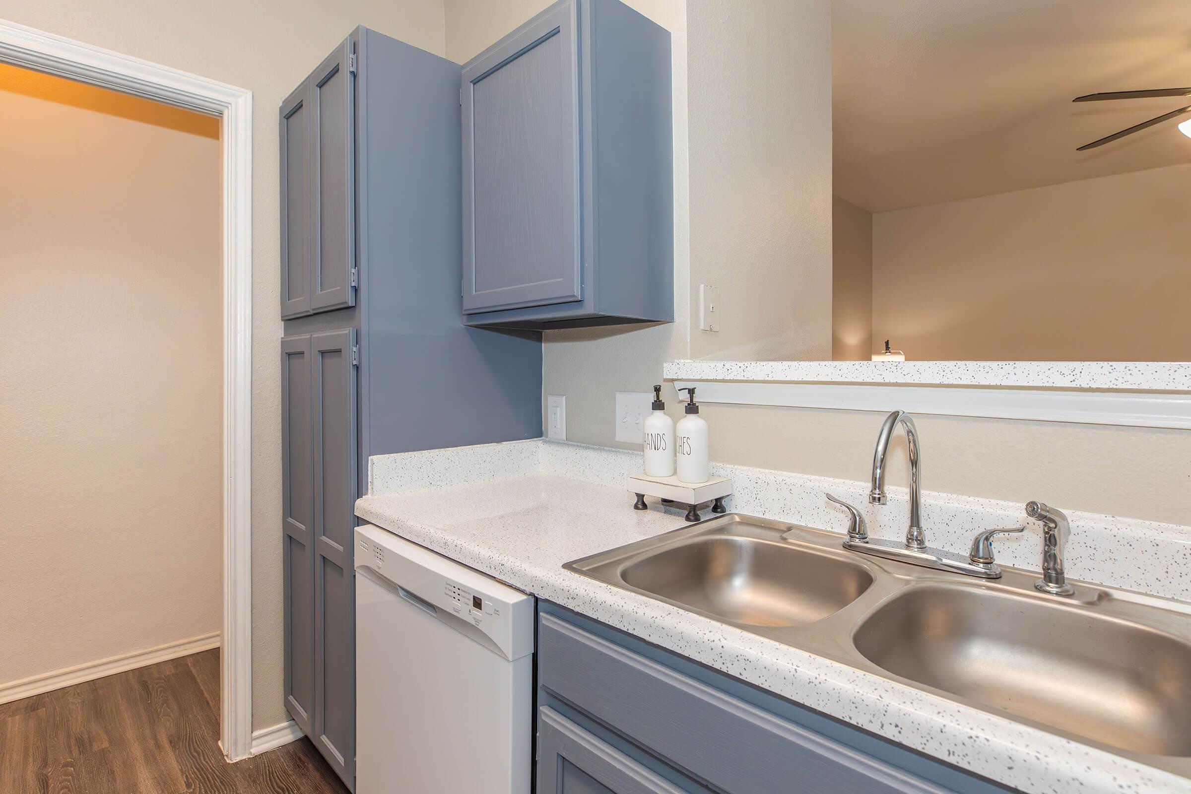 A modern kitchen featuring a gray cabinet with a white countertop, double stainless steel sinks, and a dishwasher. The space is well-lit, with a glimpse of an open area beyond. Clean and organized, the kitchen showcases a minimalist design.