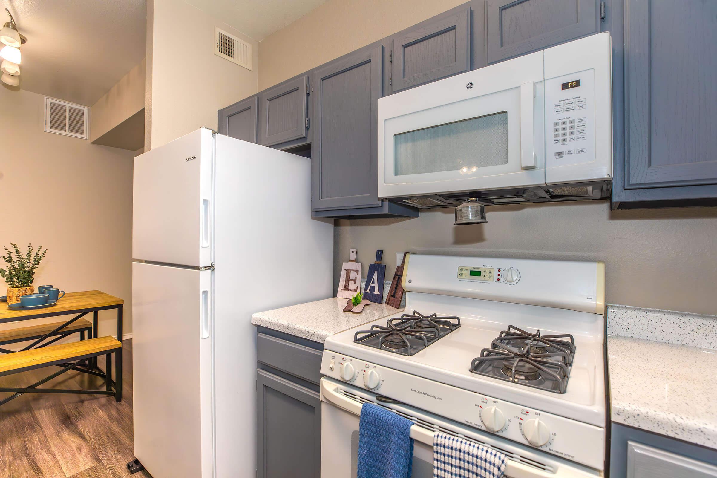 A modern kitchen featuring gray cabinets, a white refrigerator, and a gas range with a microwave above. The countertop is light-colored with decor items, and there's a small dining table with a wooden surface and black metal legs in the background.
