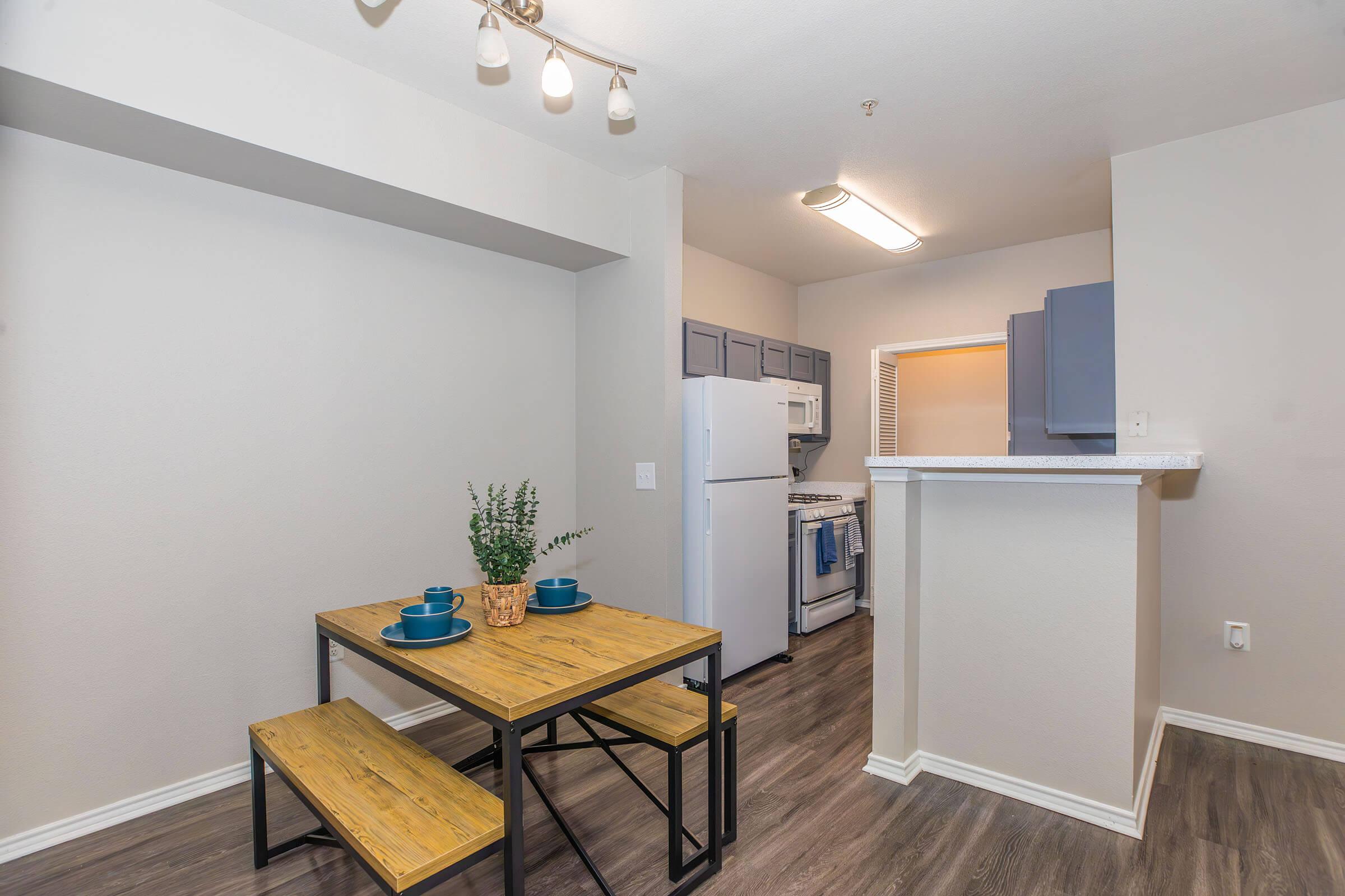 A modern kitchen and dining area featuring a wooden table with blue dishes, two benches, gray cabinets, and a white countertop. The kitchen is equipped with an oven, refrigerator, and light-colored walls, creating a bright and inviting atmosphere.