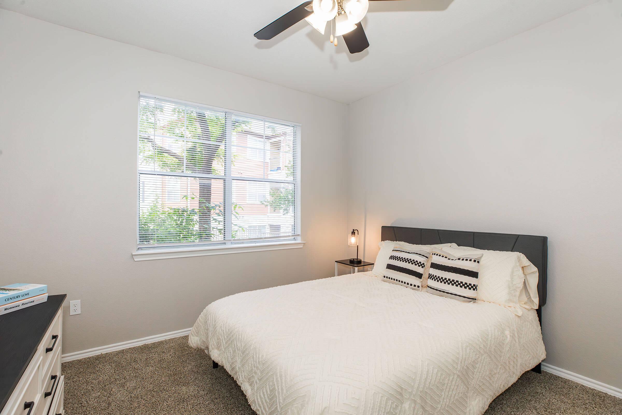 A cozy bedroom featuring a queen-sized bed with a light-colored quilt and decorative pillows. There is a nightstand with a lamp beside the bed, and a large window provides natural light, offering a view of trees outside. The room has soft beige walls and carpet, creating a warm atmosphere.