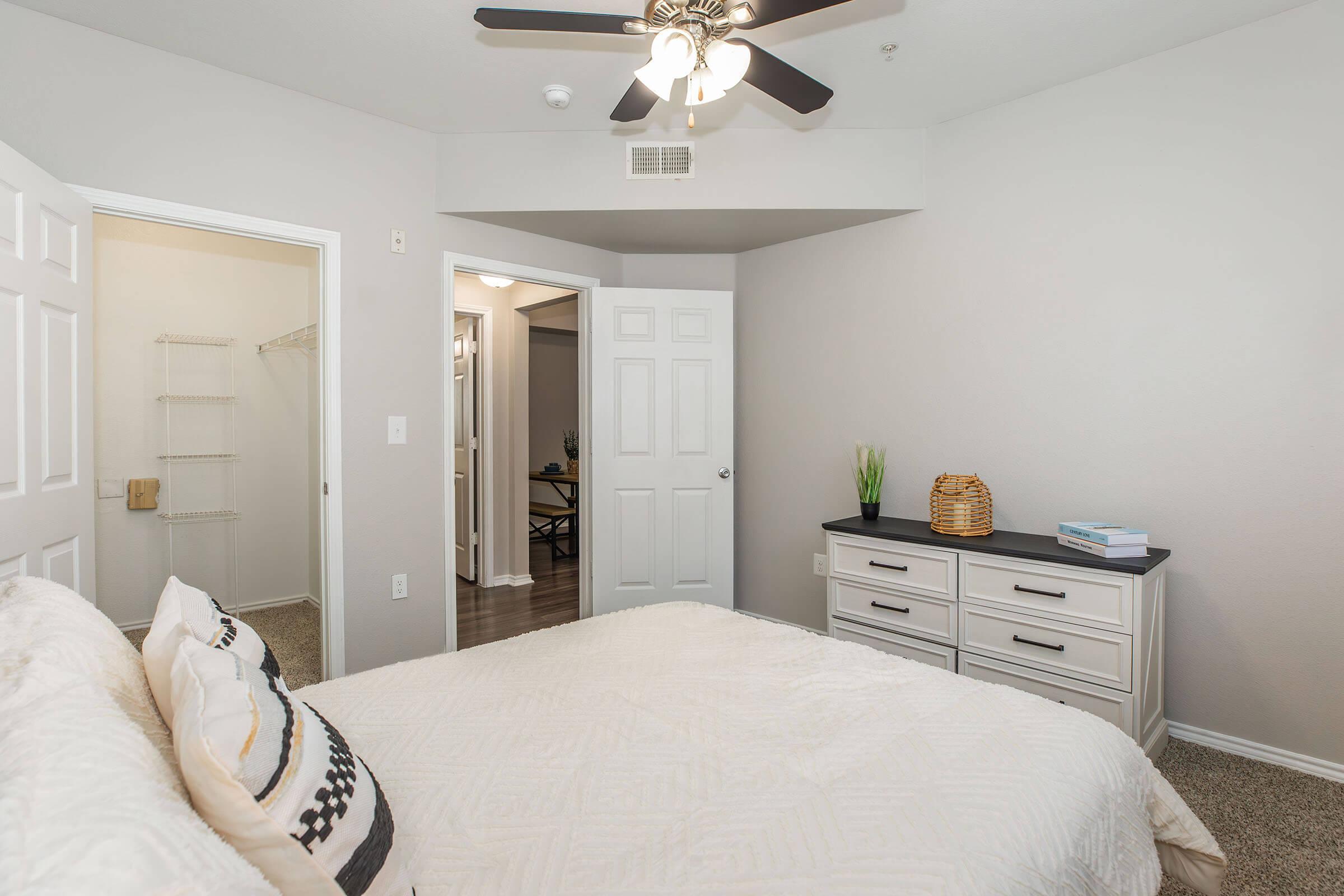A cozy bedroom featuring a light-colored wall, a queen-sized bed with decorative pillows, a dresser with a dark top, a ceiling fan, and a door leading to another room. A closet is visible on the left, and a small green plant adds a touch of decor. The overall atmosphere is bright and inviting.