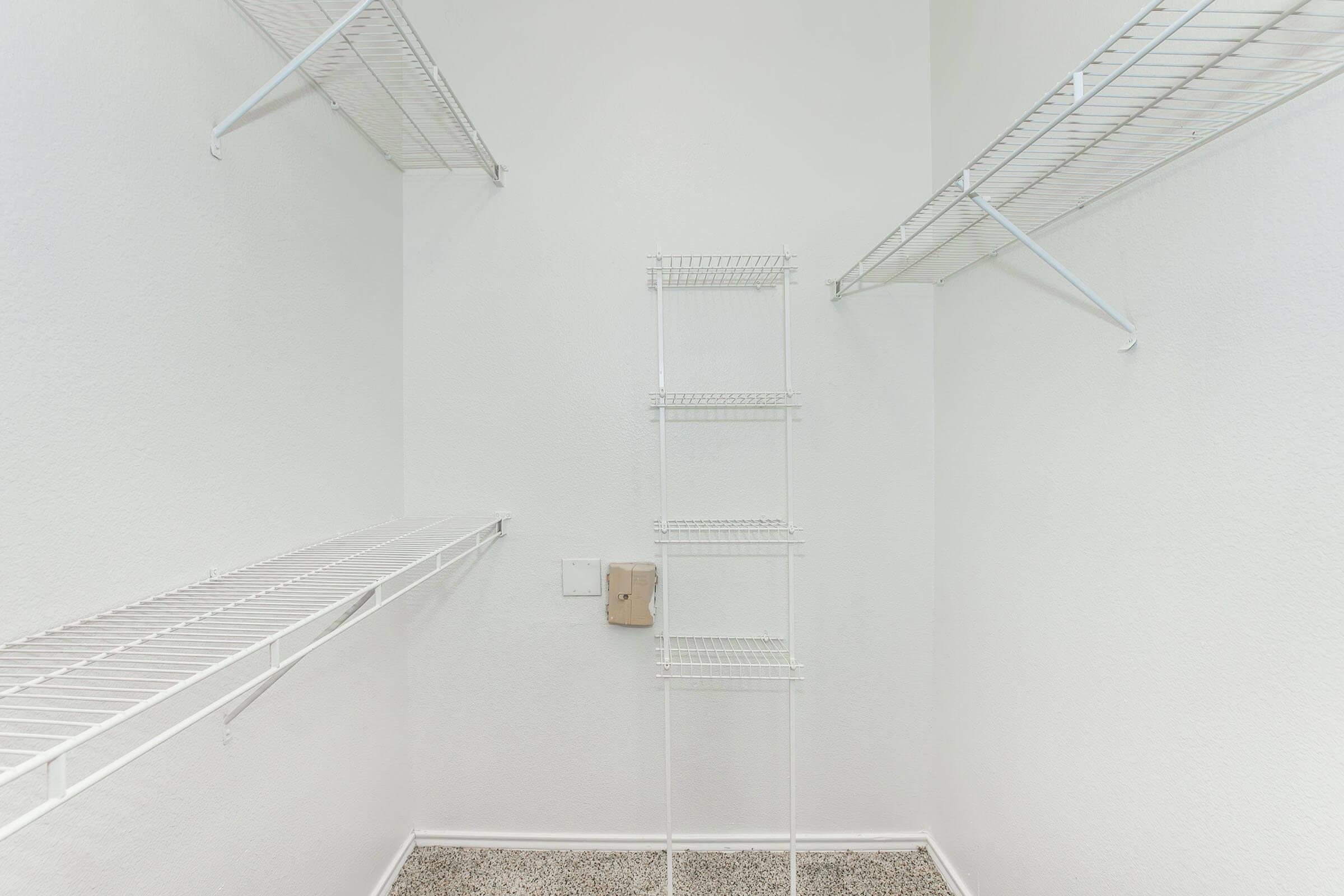 An empty walk-in closet featuring white wire shelving on the walls, designed for storage. The walls are painted white, and the floor is carpeted in a neutral color. There's a small wire rack in the center, standing alone without any items on display. The overall look is clean and organized.