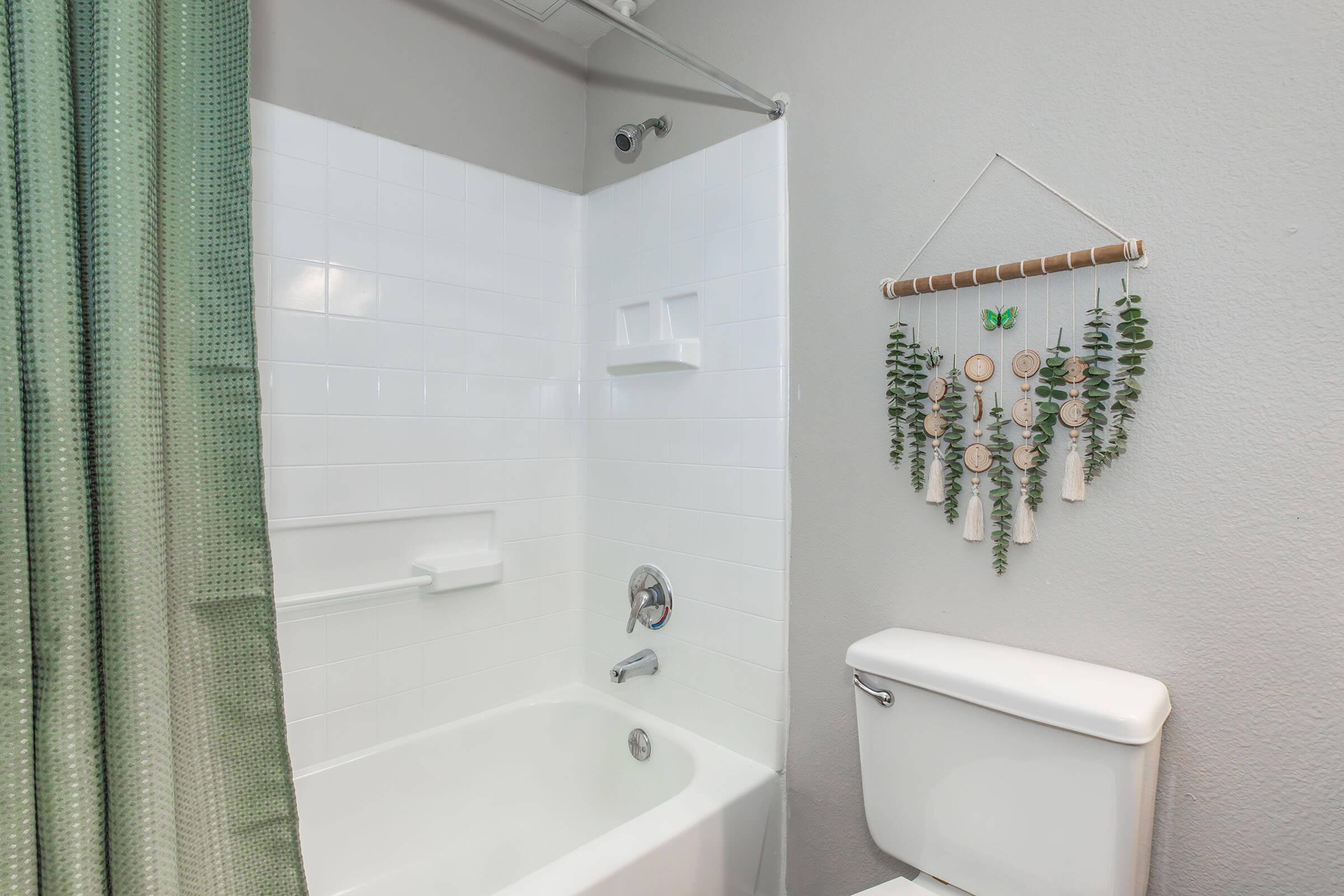 A clean bathroom featuring a white bathtub with a shower curtain in green, a white toilet, and a decorative wall hanging with natural elements. The walls are painted light gray, and there are simple shelves for toiletries. Natural light brightens the space.