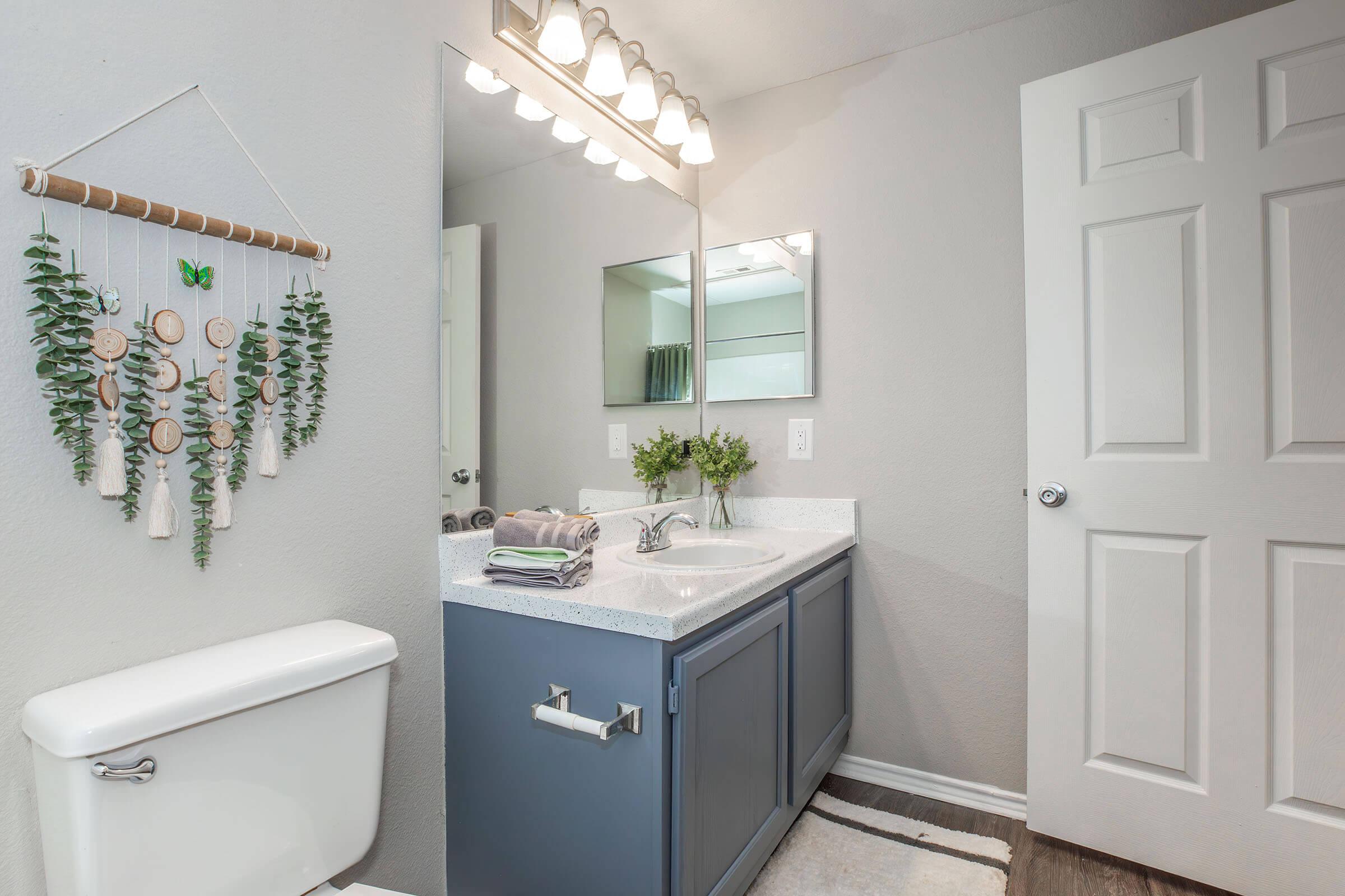 Modern bathroom featuring a rectangular vanity with a white countertop and blue cabinetry, a large mirror above the sink, and decorative plants. To the left, there is a white toilet, and on the wall, a hanging display of wooden circles with greenery. A bright light fixture illuminates the space.