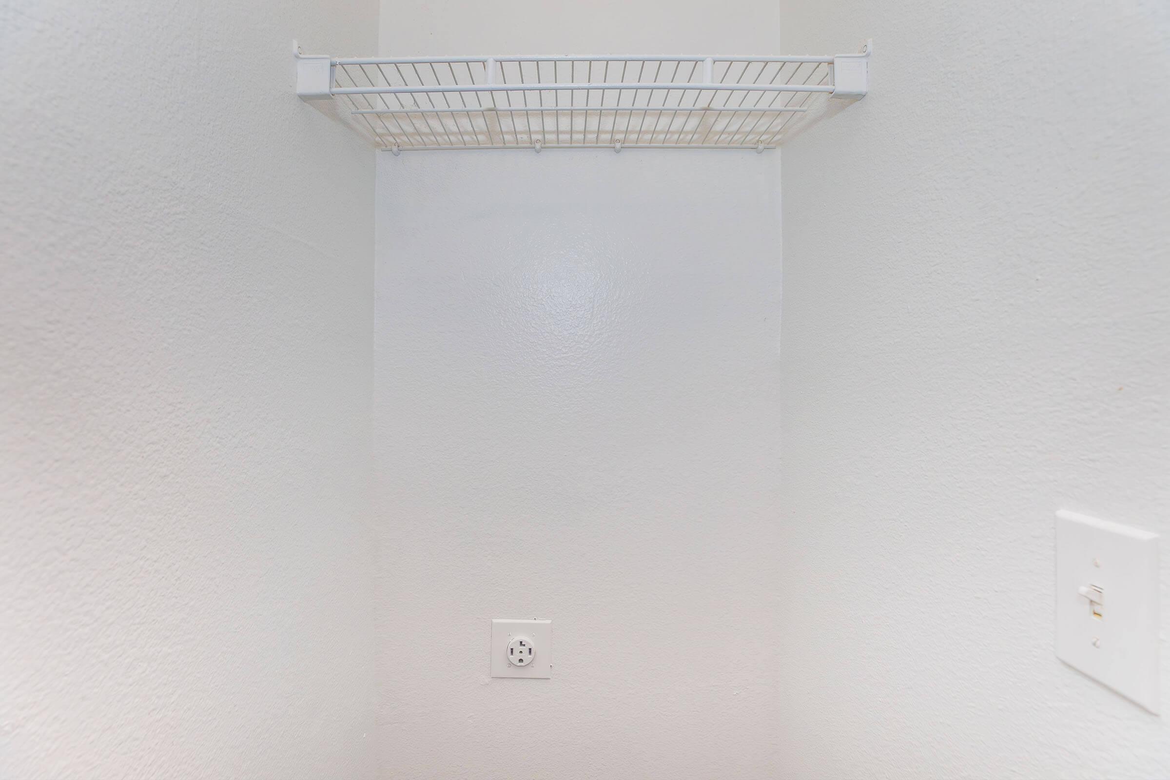 Empty closet corner with a white wall and a wire shelf attached to the upper wall. There is a power outlet on the right side of the image. The overall space appears clean and minimalistic.