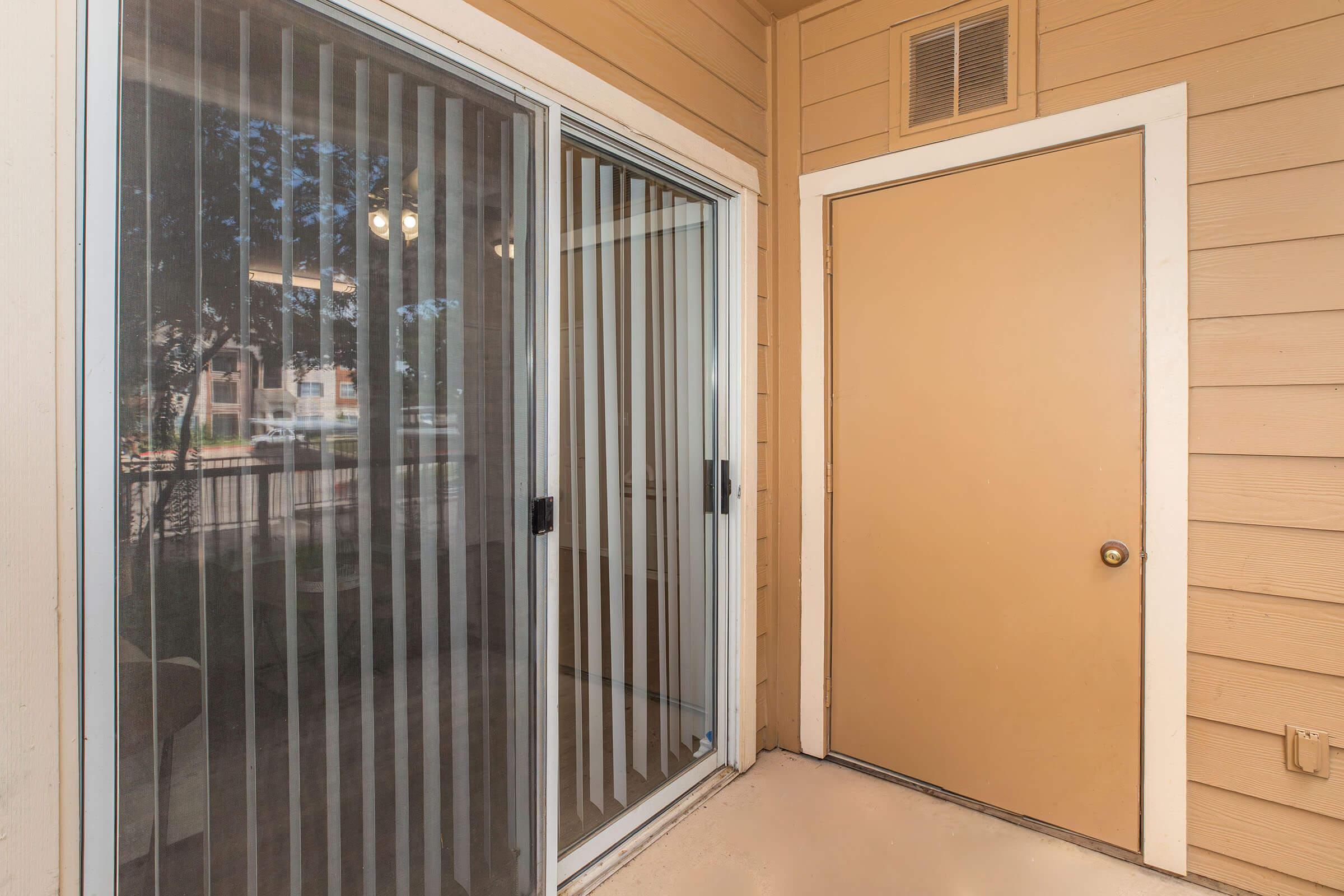 A patio area featuring a sliding glass door with vertical blinds and a solid brown door. The walls are painted a warm beige color, and the floor appears to be a light-colored concrete. Natural light filters through the blinds, illuminating the space.