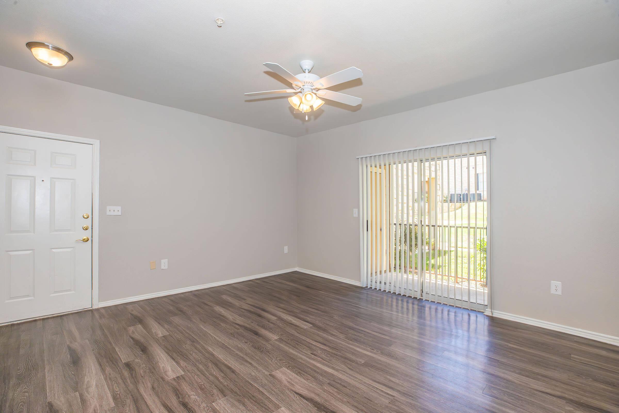 A spacious and well-lit living room featuring laminate flooring, a ceiling fan, and a sliding glass door with vertical blinds that leads to an outdoor area. The walls are painted in a light color, enhancing the airy atmosphere. A front door is visible on the left side of the room.