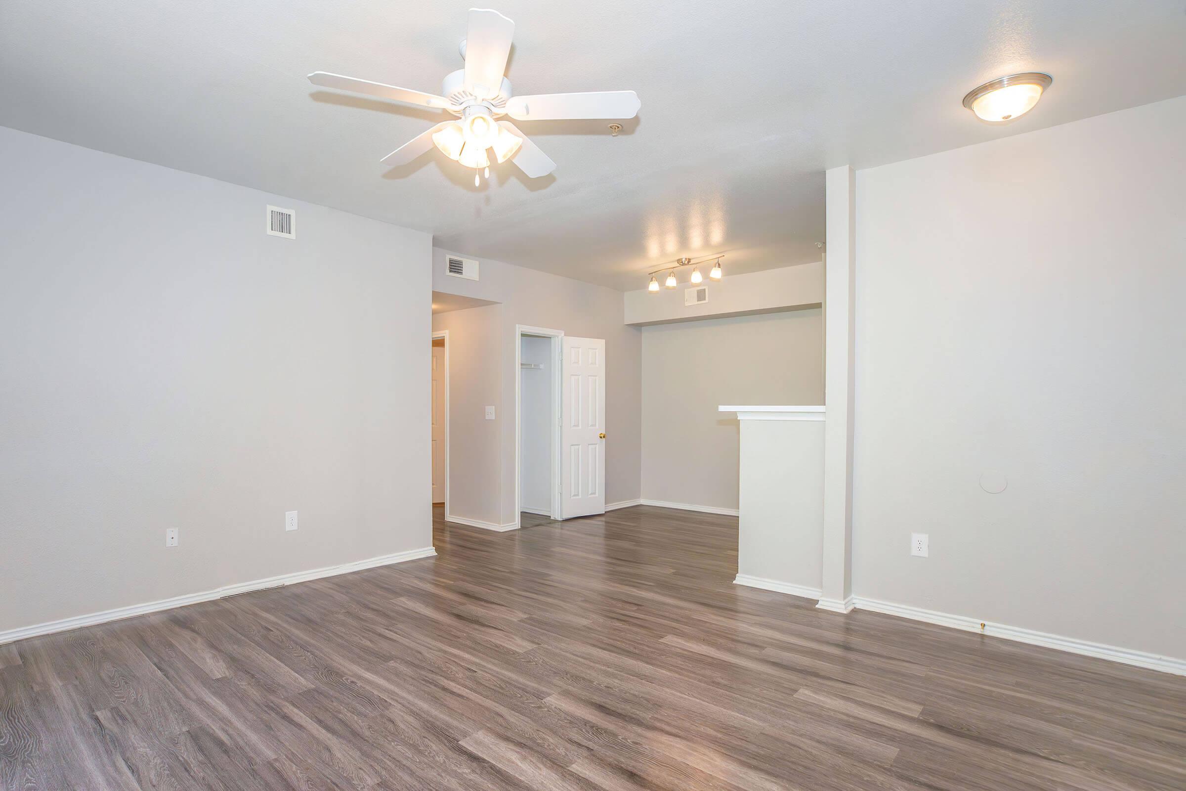 A spacious living area featuring a ceiling fan, light gray walls, and laminate flooring. There are two doorways leading to other rooms, and a small wall partition. The space is well-lit with natural light, creating a welcoming atmosphere.