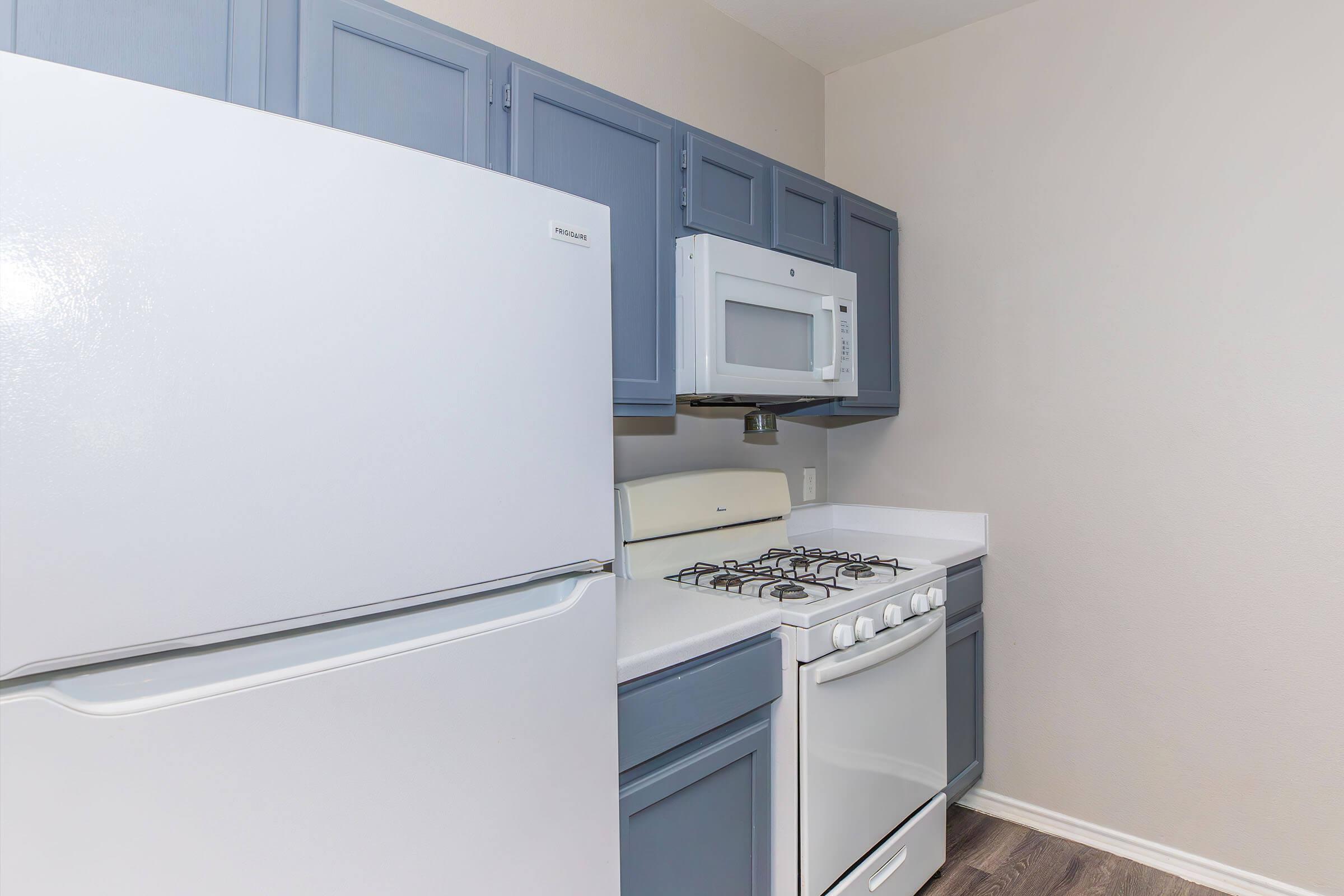 A modern kitchen featuring a white refrigerator, a microwave above a white stove with a gas cooktop, and blue cabinets. The countertops are white, and the flooring is dark wood. The walls are a light color, creating a bright and airy atmosphere.