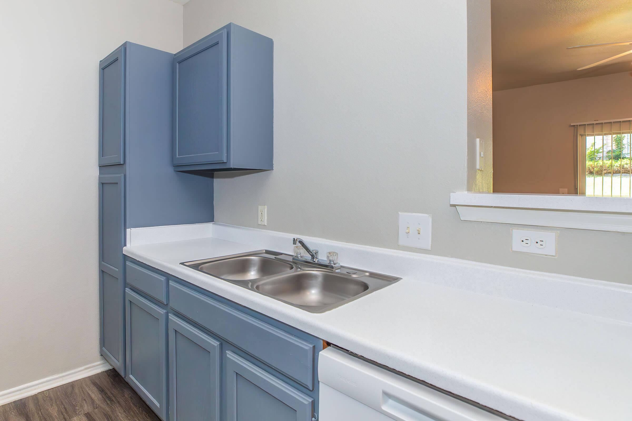 A modern kitchen featuring gray cabinets, a double sink with chrome faucets, and a white countertop. There's a dishwasher visible in the lower section, and the scene is well-lit, with a view into a bright, airy space beyond.