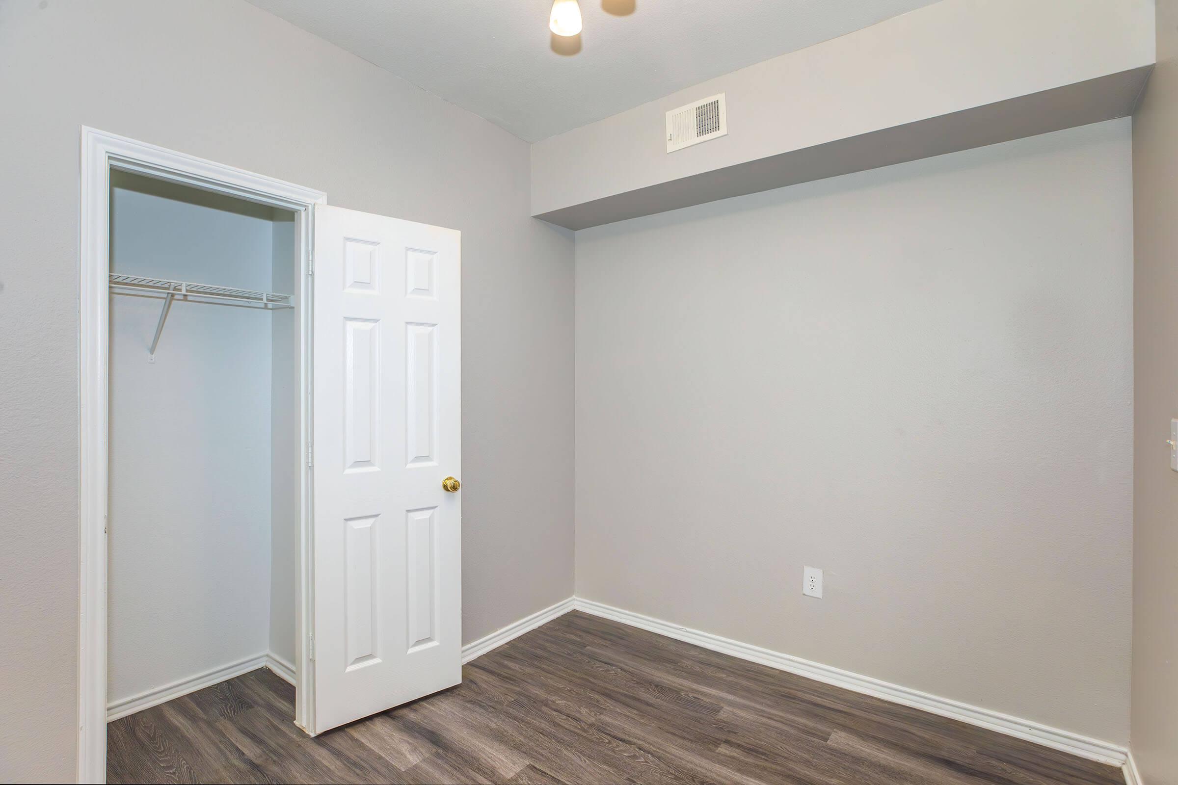 A small, empty room with light gray walls and a wooden floor. There is a white closet door on the left, leading to an empty closet, and a ceiling fan with a light fixture above. The room has a clean, minimalist appearance, suitable for various uses.