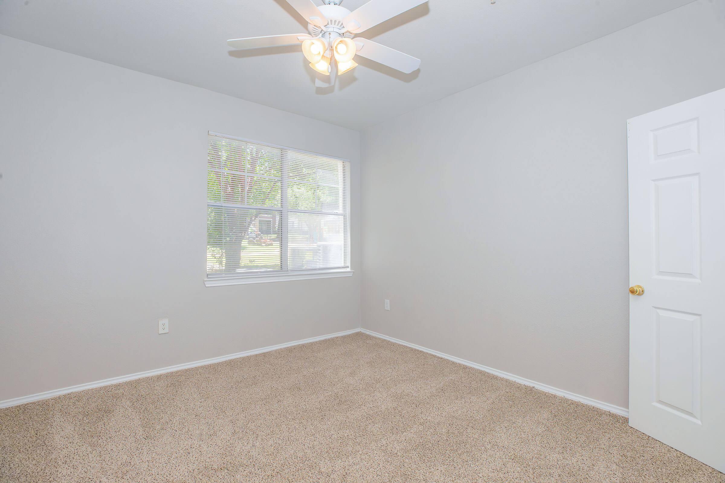 Empty room with light gray walls and beige carpet. A ceiling fan with light fixtures is mounted in the center. A window with blinds allows natural light to enter, and a white door is visible on the right side of the image. The space appears clean and ready for furnishing.