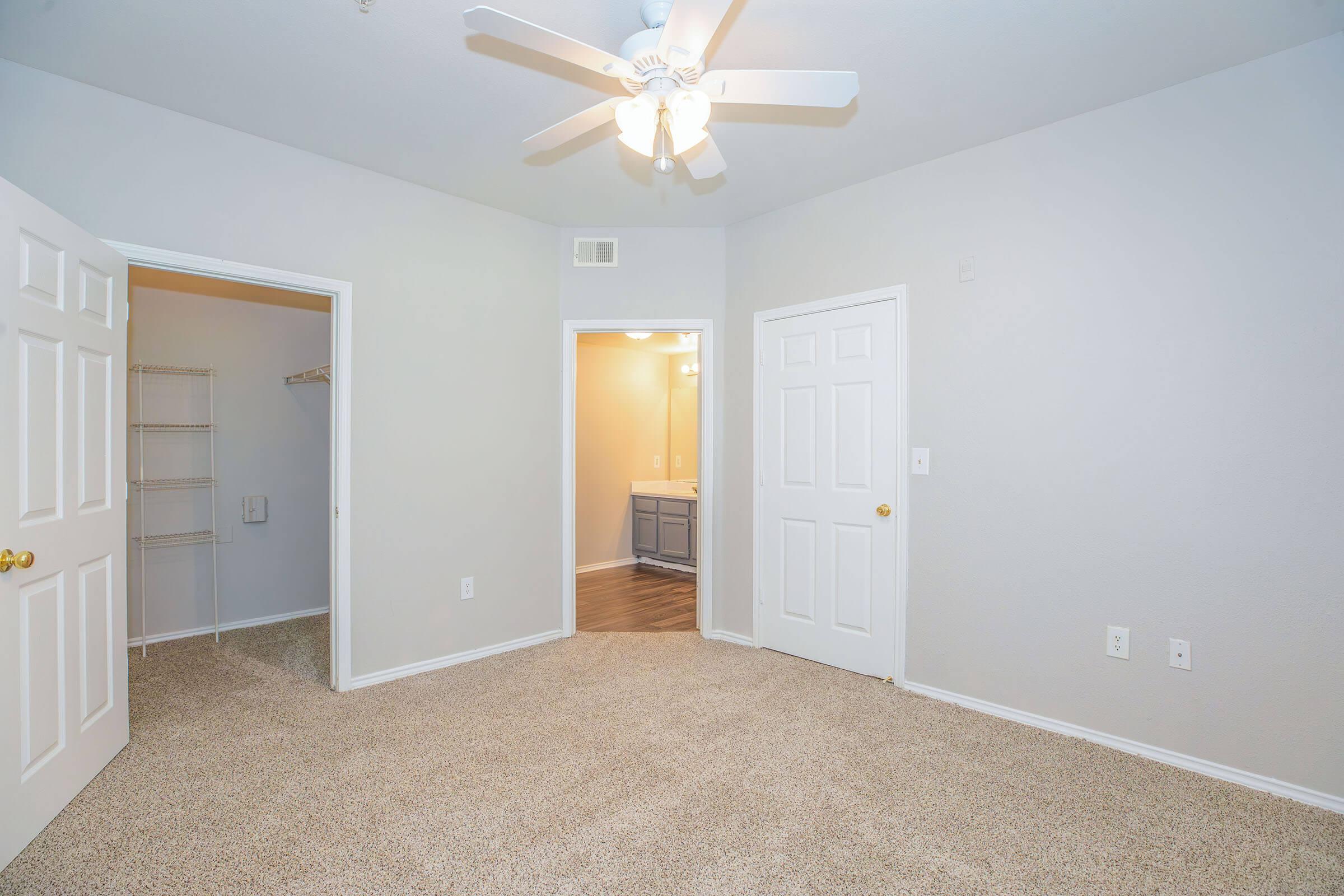 A well-lit room featuring light gray walls, a ceiling fan, and plush beige carpet. Two white doors lead to a closet and a bathroom area, respectively. The space appears clean and inviting, suitable for various furnishings and decor.