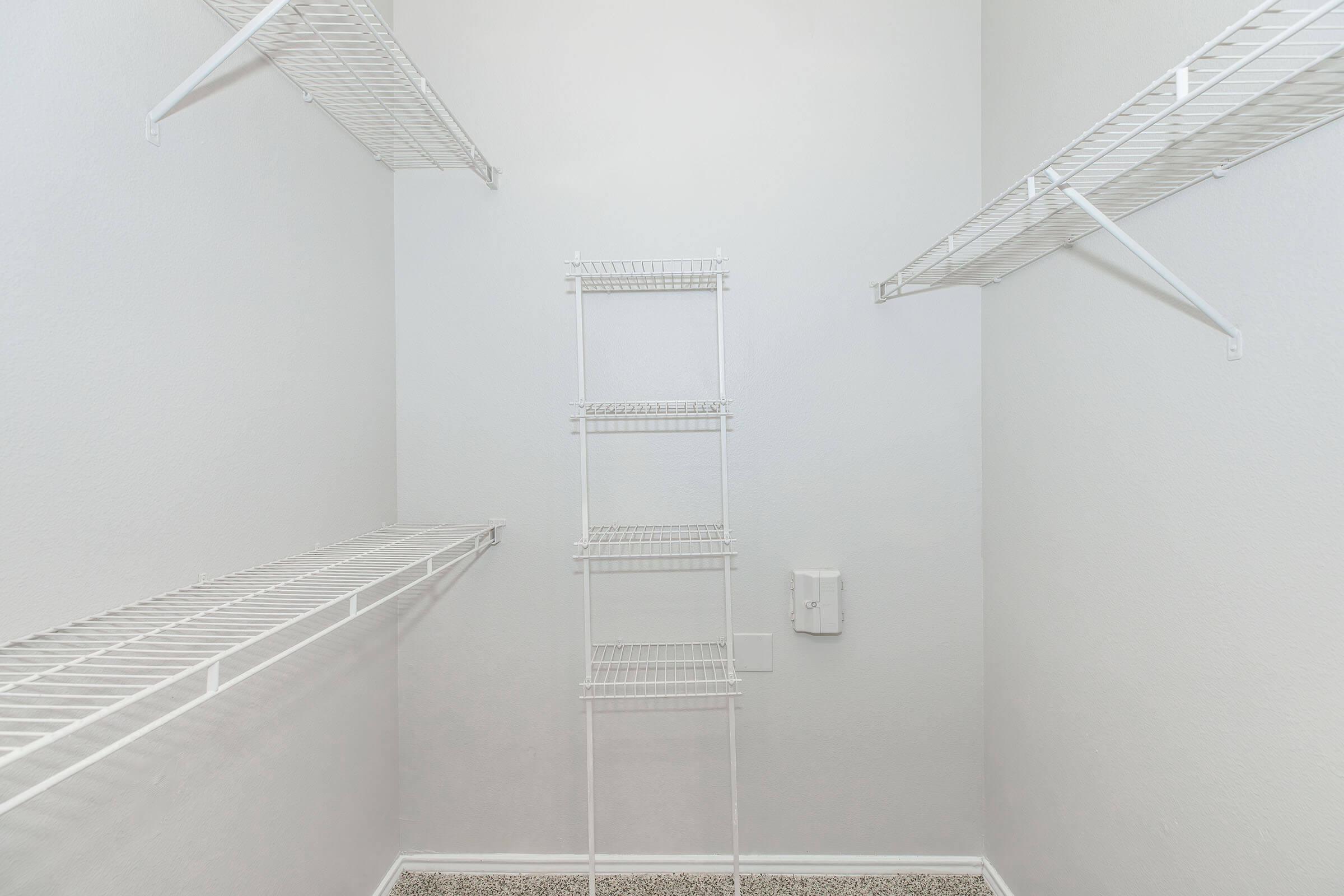 A spacious, empty closet featuring white wire shelving on the walls and a standalone wire shelf in the center. The walls are painted a light color, and the carpeted floor is visible. The overall appearance is clean and organized, providing ample storage space.