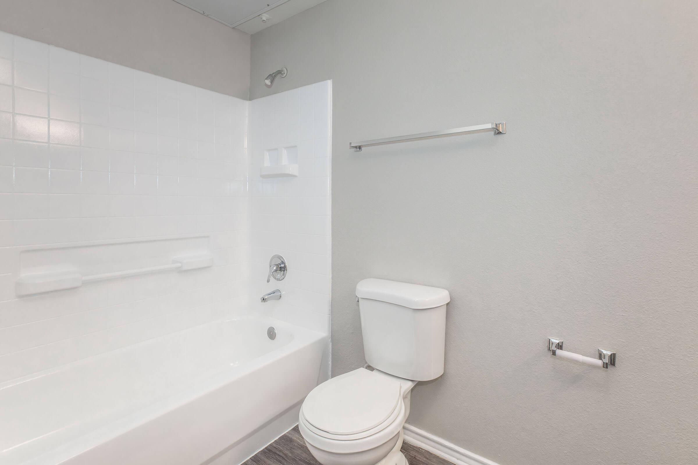 A clean, modern bathroom featuring a white bathtub and shower combination, a white toilet, and a light gray wall. There is a grab bar for safety, a towel bar, and a simple design that emphasizes a spacious and uncluttered appearance. The flooring has a wood-like texture.