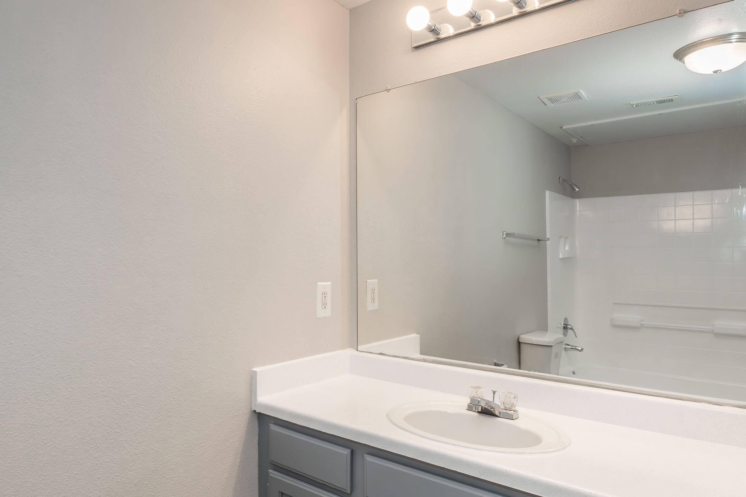 A clean, modern bathroom featuring a light-colored wall, a large mirror, and a white sink with a faucet. The countertop is smooth and simple, while the background shows a shower area with white tiles and a toilet. Natural and artificial lighting enhances the bright atmosphere of the space.