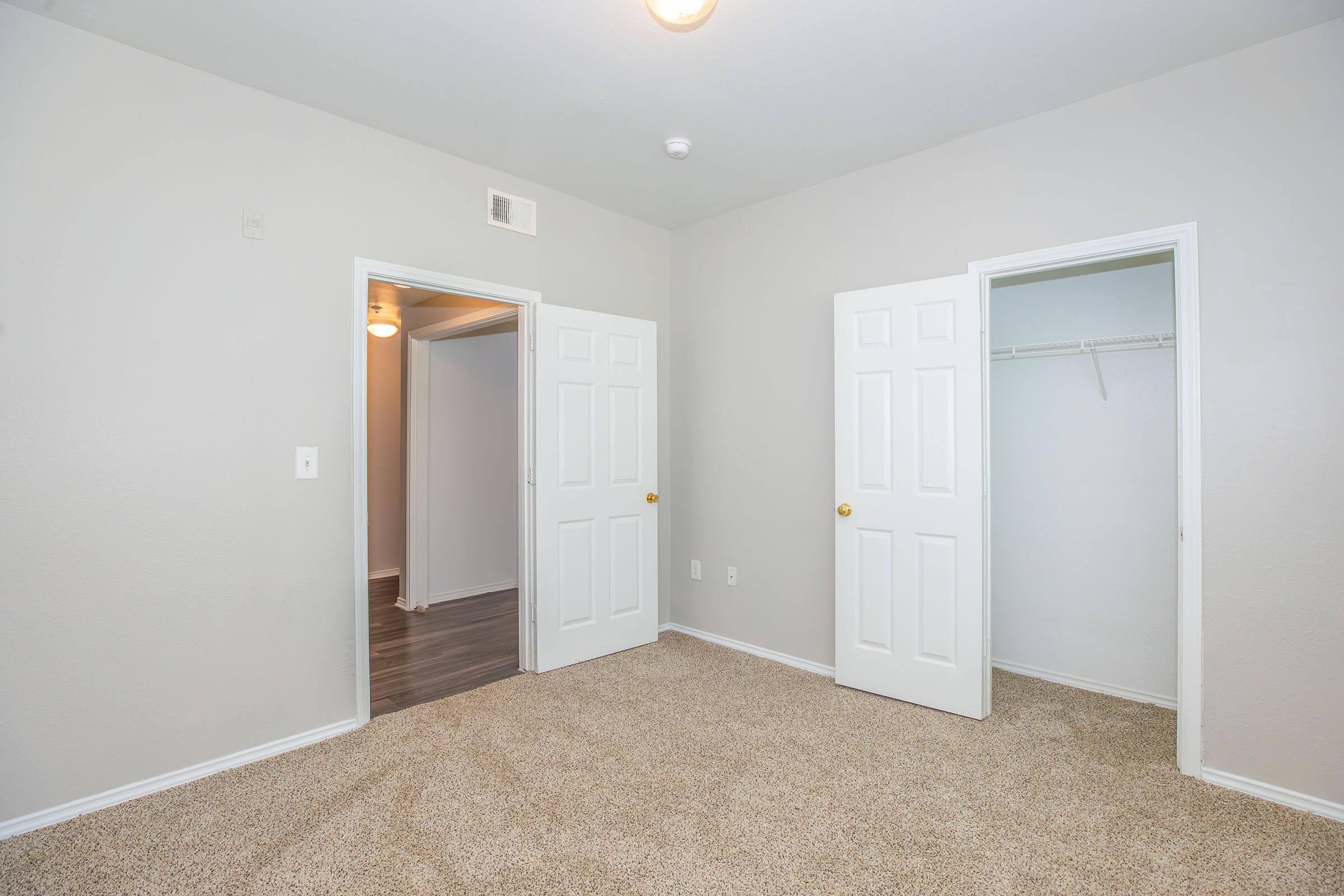 Empty room with light gray walls and carpeted flooring. Two white doors are visible: one leading to a closet and the other to another space. A ceiling light fixture illuminates the room, which has no furniture or decorations.