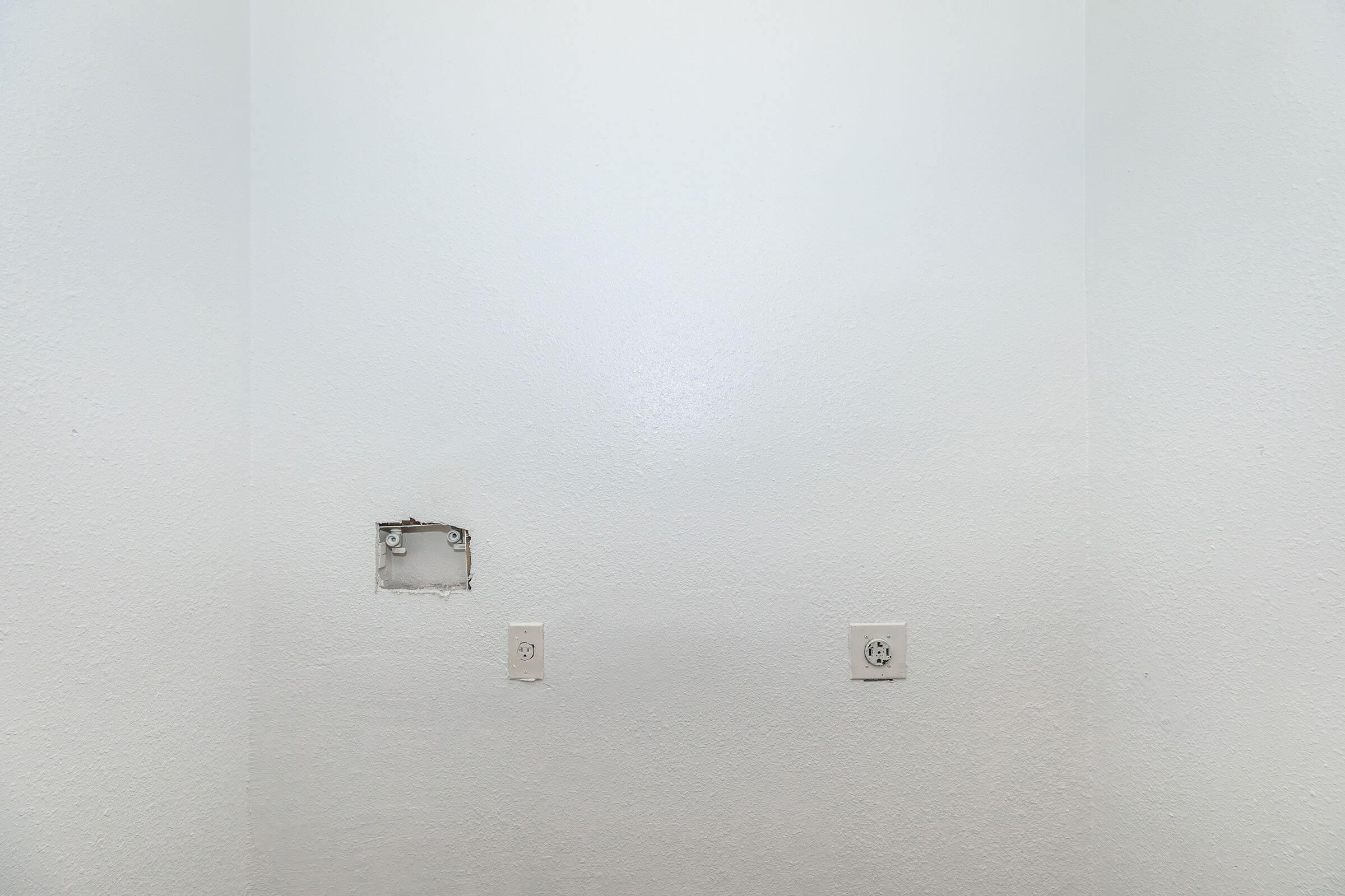 A plain white wall featuring two electrical outlets and a small rectangular cutout that appears to be an access panel. The surface has a subtle texture, with no other objects or decorations present.
