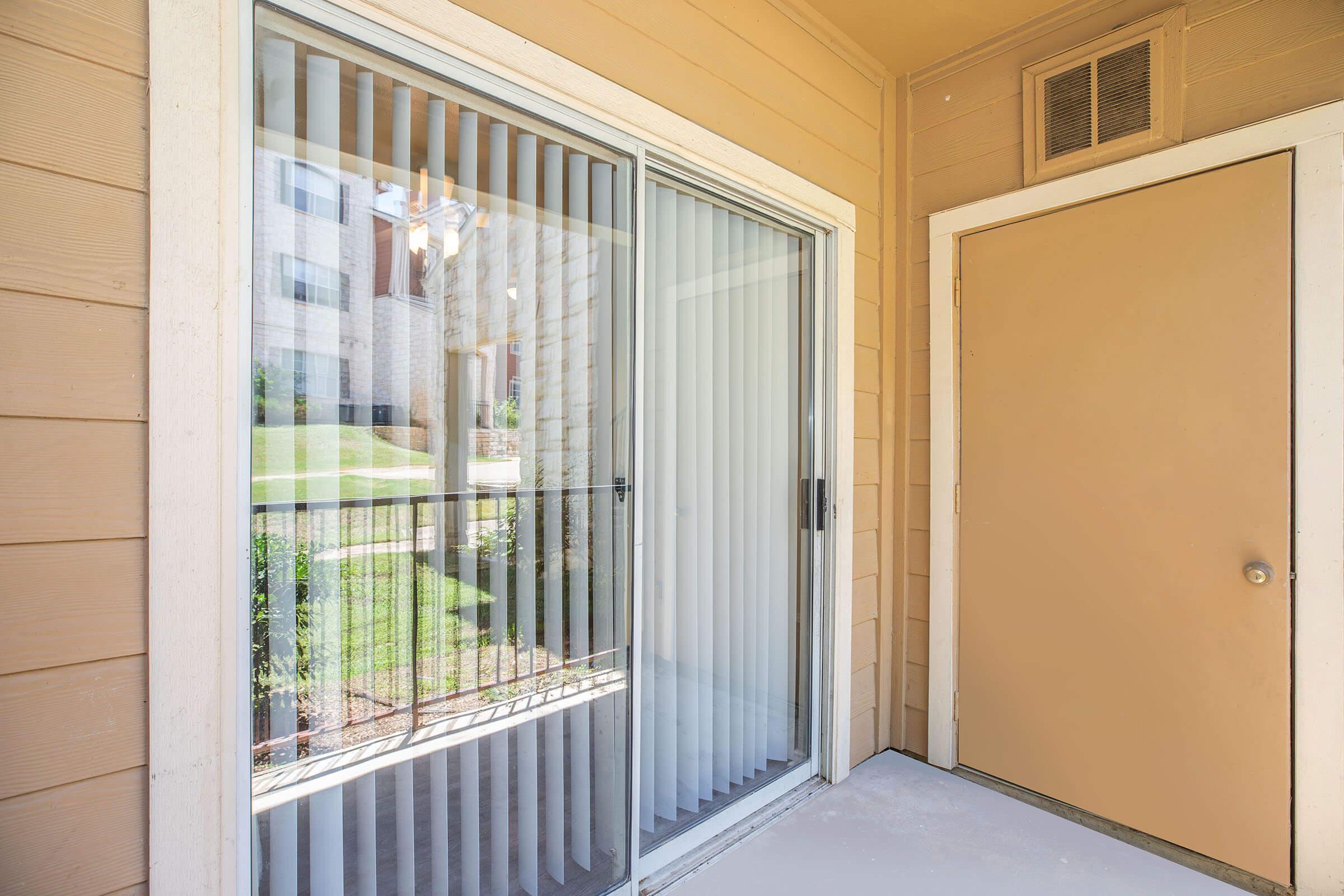 A patio area featuring a sliding glass door with vertical blinds, leading to a grassy outdoor space. There is a solid-colored door on the right side, matching the beige exterior. The wall has a textured finish, and the setting is bright and inviting.